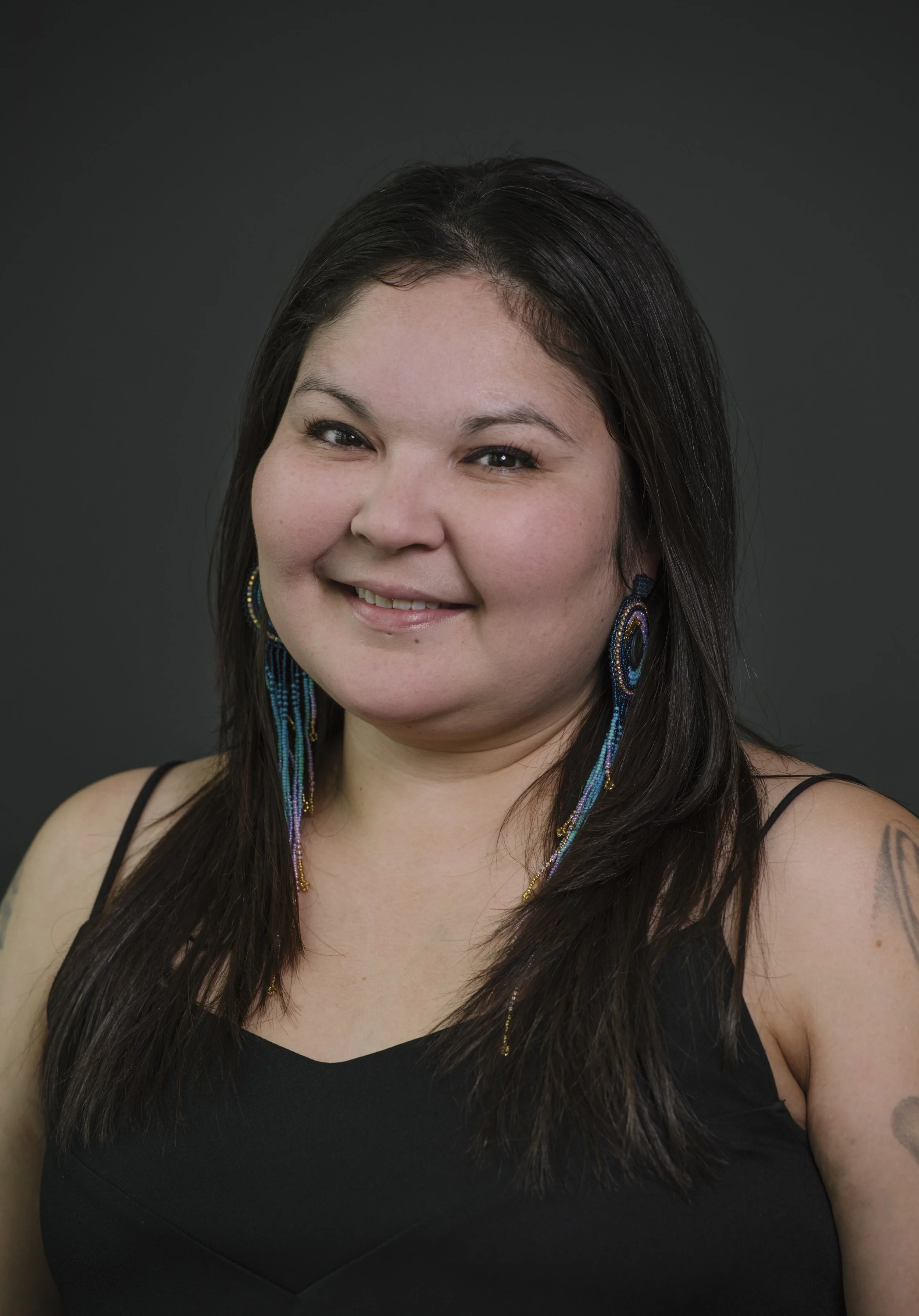Portrait of a woman with long dark hair, wearing colorful beaded earrings and a black sleeveless top, smiling against a dark background.