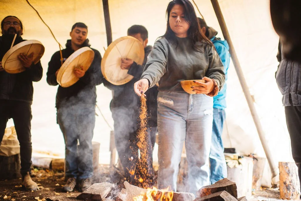 A woman sprinkling what appears to be ash or spices over an open campfire inside a tent, with several people in the background playing drums.