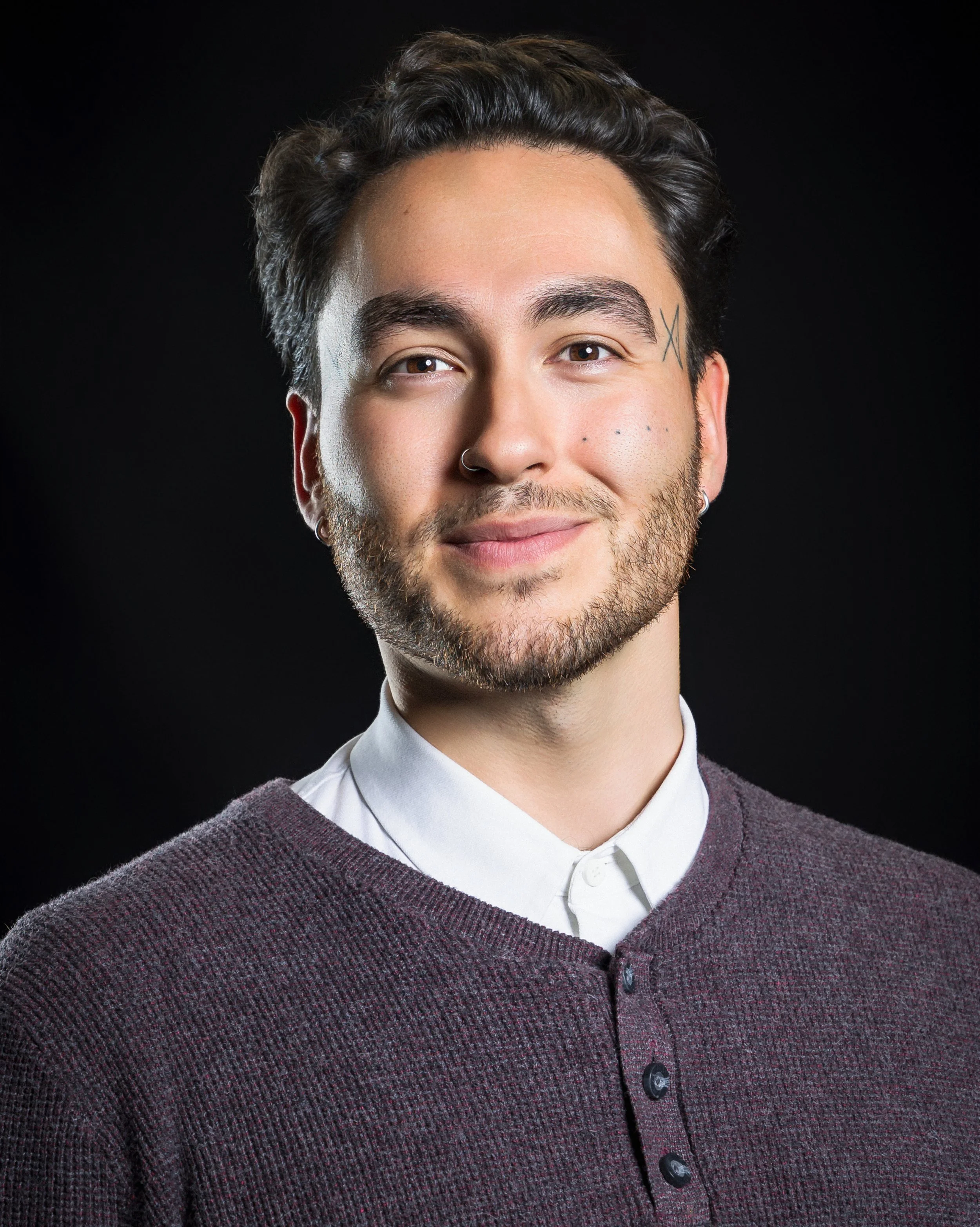 A young man with dark hair and beard, wearing a white shirt and a brown sweater, smiling against a black background.
