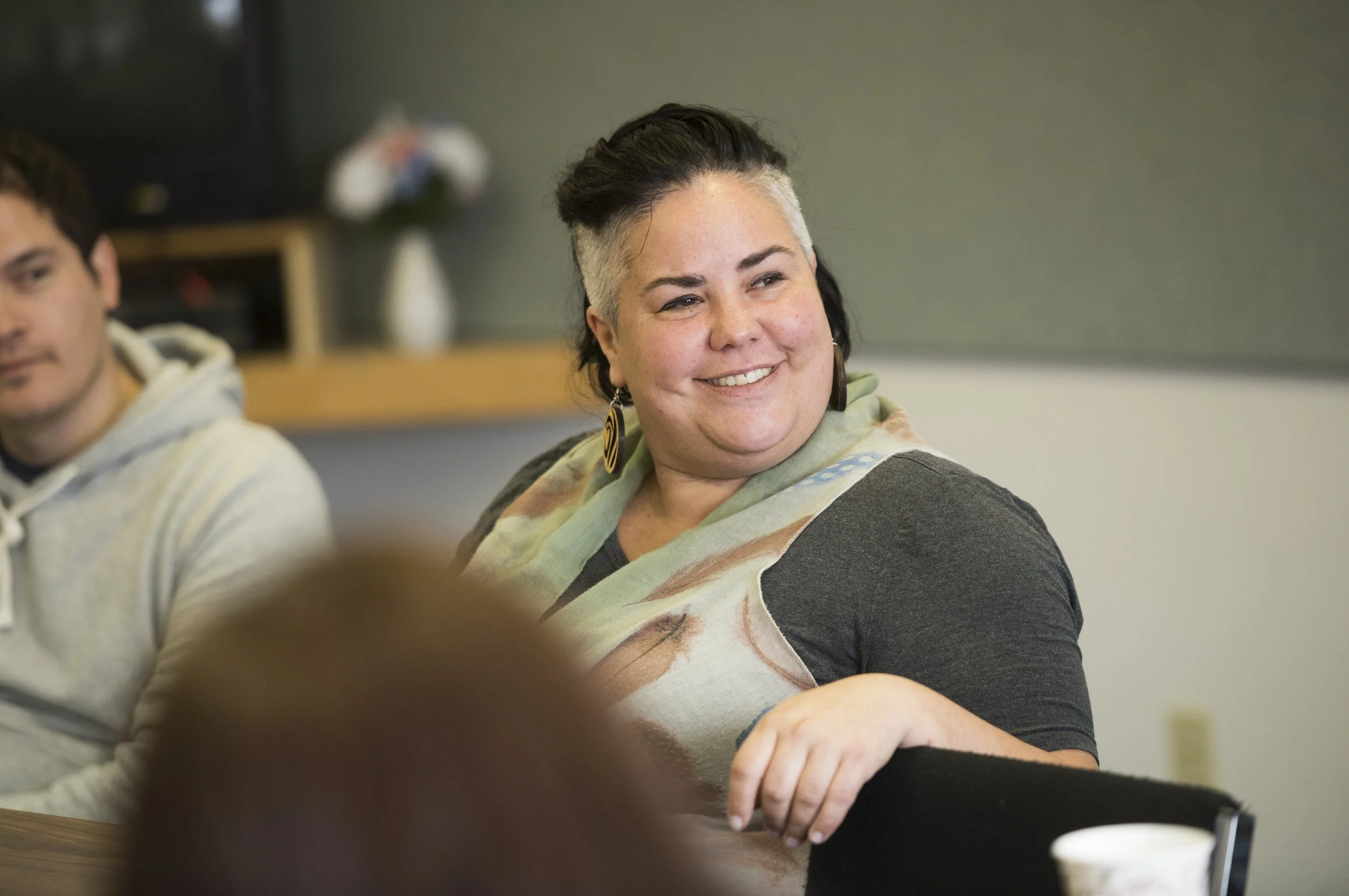 A smiling woman with short, dark hair and gray sides, wearing a colorful scarf and earrings, sits at a table in a room with a computer monitor and a blackboard in the background.