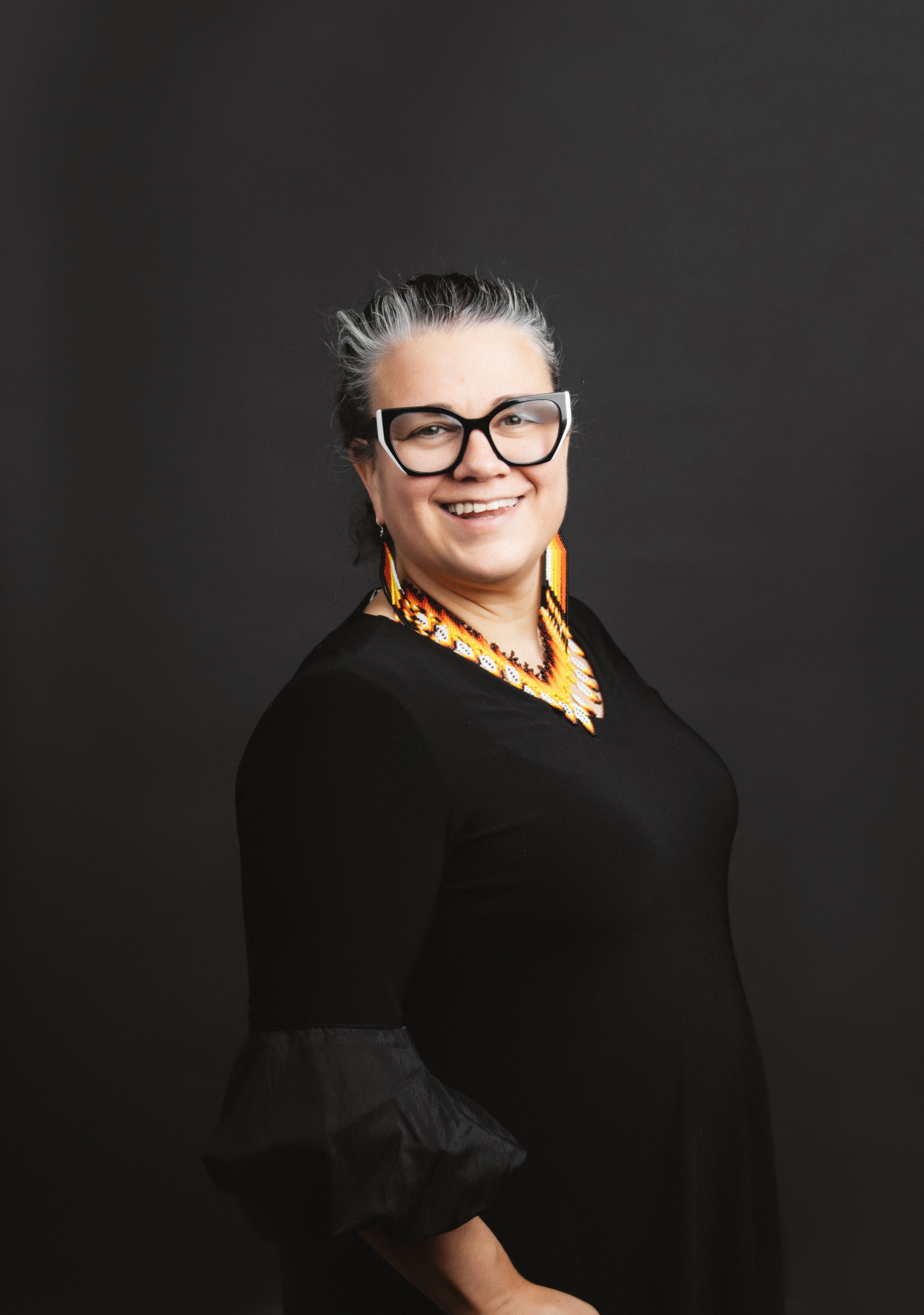 A woman with glasses, jewelry, and a black dress smiling against a dark background.