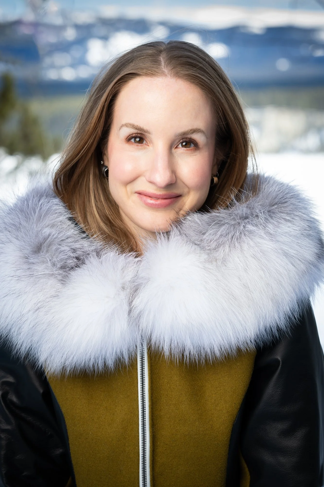 A woman smiling outdoors in winter, wearing a black winter coat with a large gray and white fur collar, snow and mountains in the background.