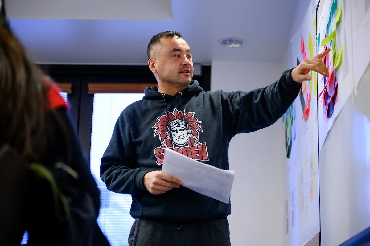A man wearing a black hoodie with a graphic of a Native American chief and the word "SACDEN", pointing at a whiteboard covered with colorful sticky notes while holding papers.