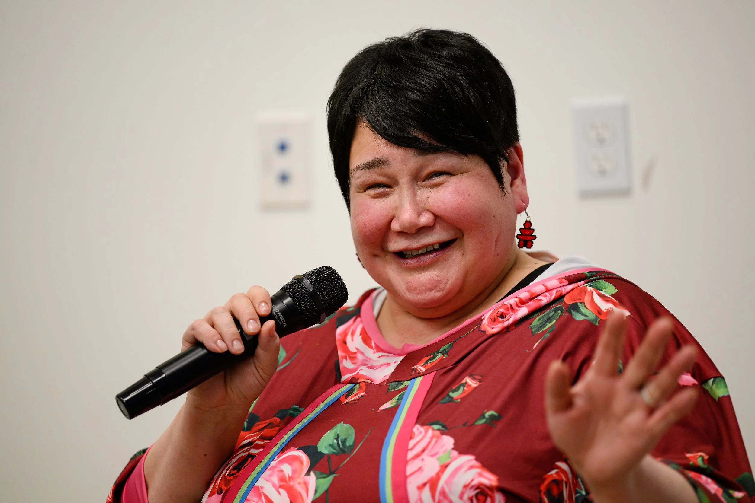 A woman with short black hair smiling and holding a microphone, wearing a red floral dress with pink and green accents, and red earrings shaped like pixelated hearts.