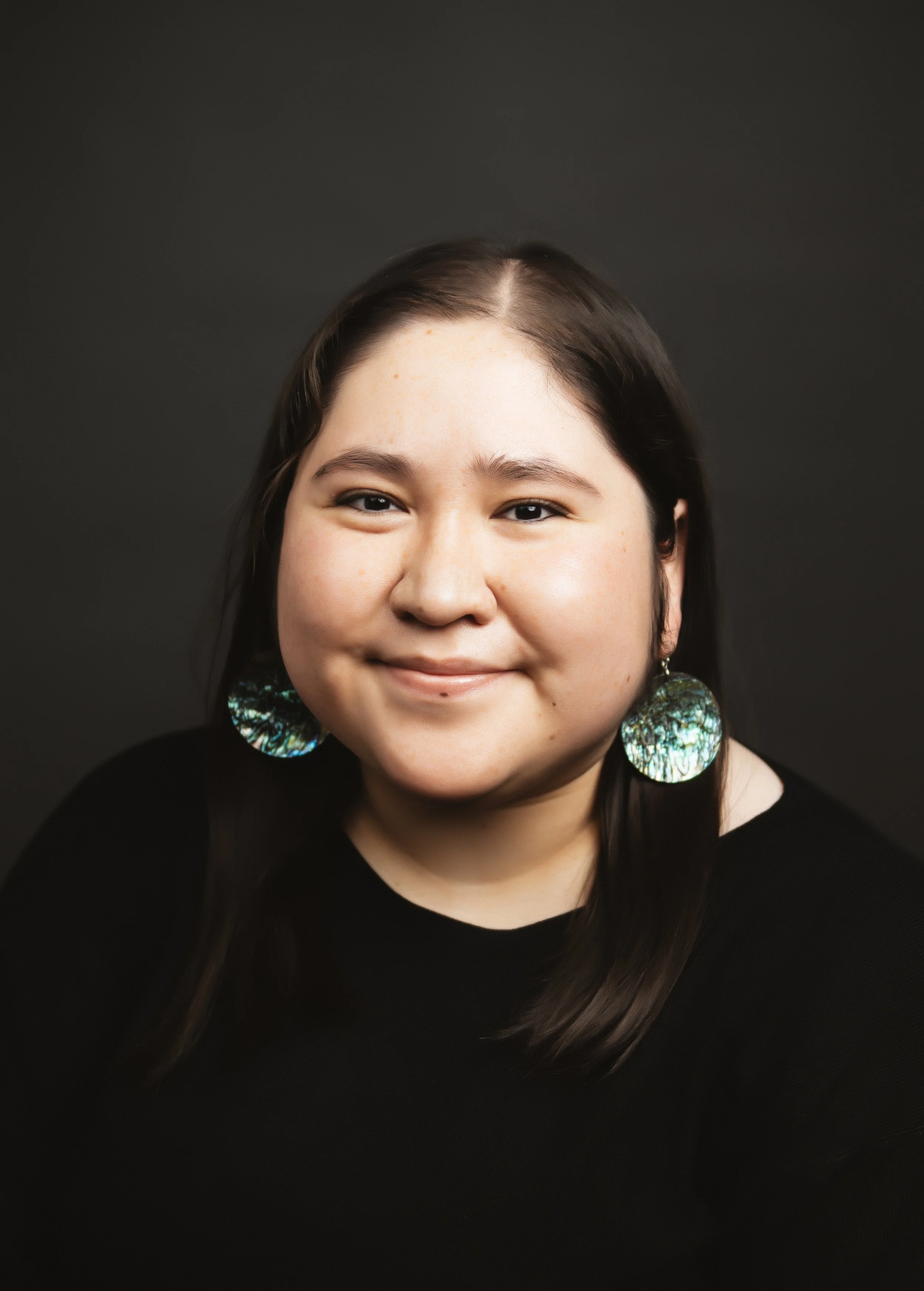 A young woman with medium-length dark hair, smiling, wearing large iridescent earrings and a black top, against a dark background.