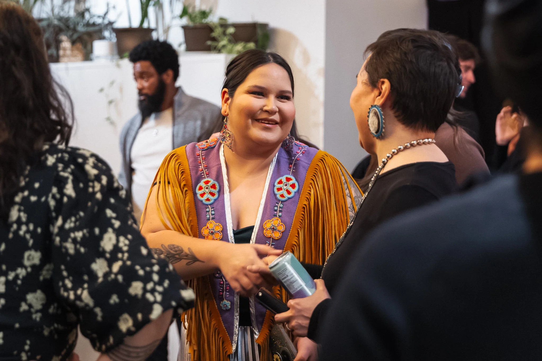 Group of people talking at an indoor gathering, with a woman in the center wearing a colorful, fringed shawl and large earrings, smiling and engaging with others around her.