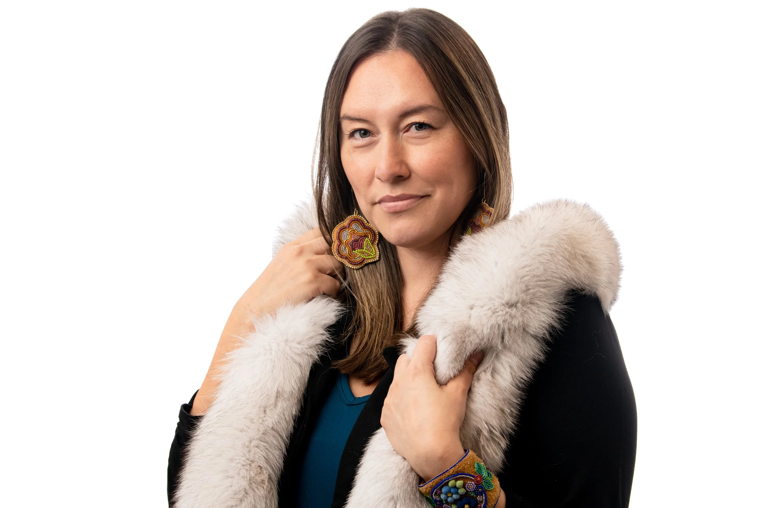 Woman with long brown hair wearing beaded earrings and a fur-lined black coat, posing against a white background.