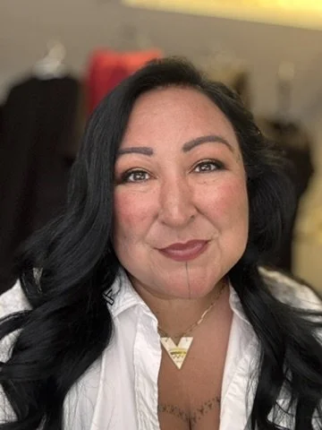 A woman with long black hair, wearing a white shirt and gold jewelry, smiling at the camera.