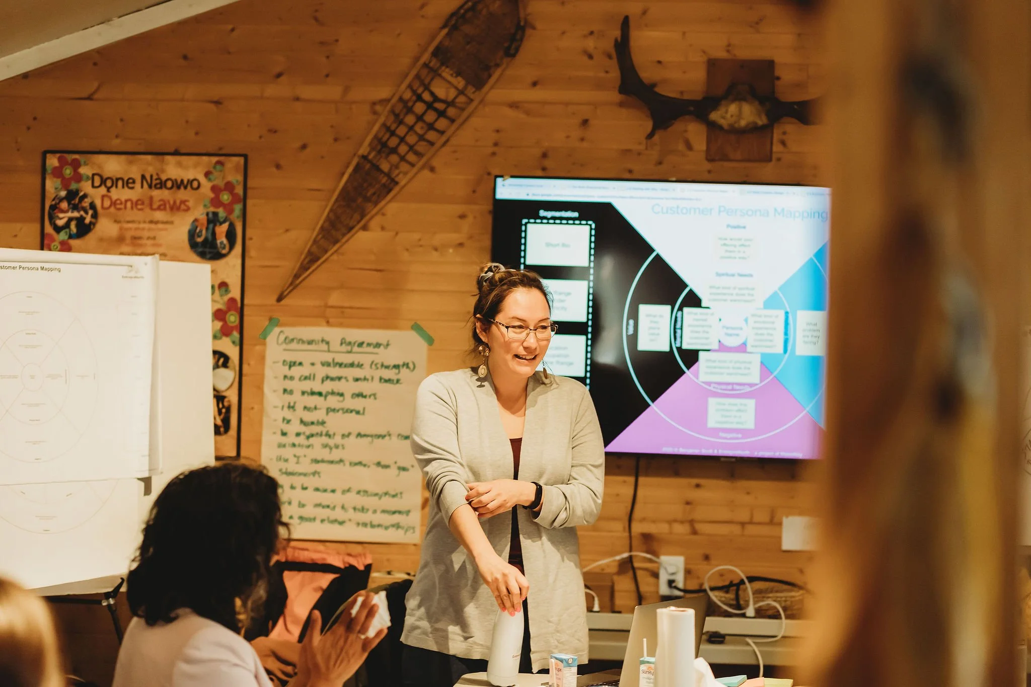 A woman smiling and speaking in front of a presentation screen displaying a customer persona mapping diagram, with another person clapping nearby in a wooden-paneled room with snowshoe and moose head decorations.