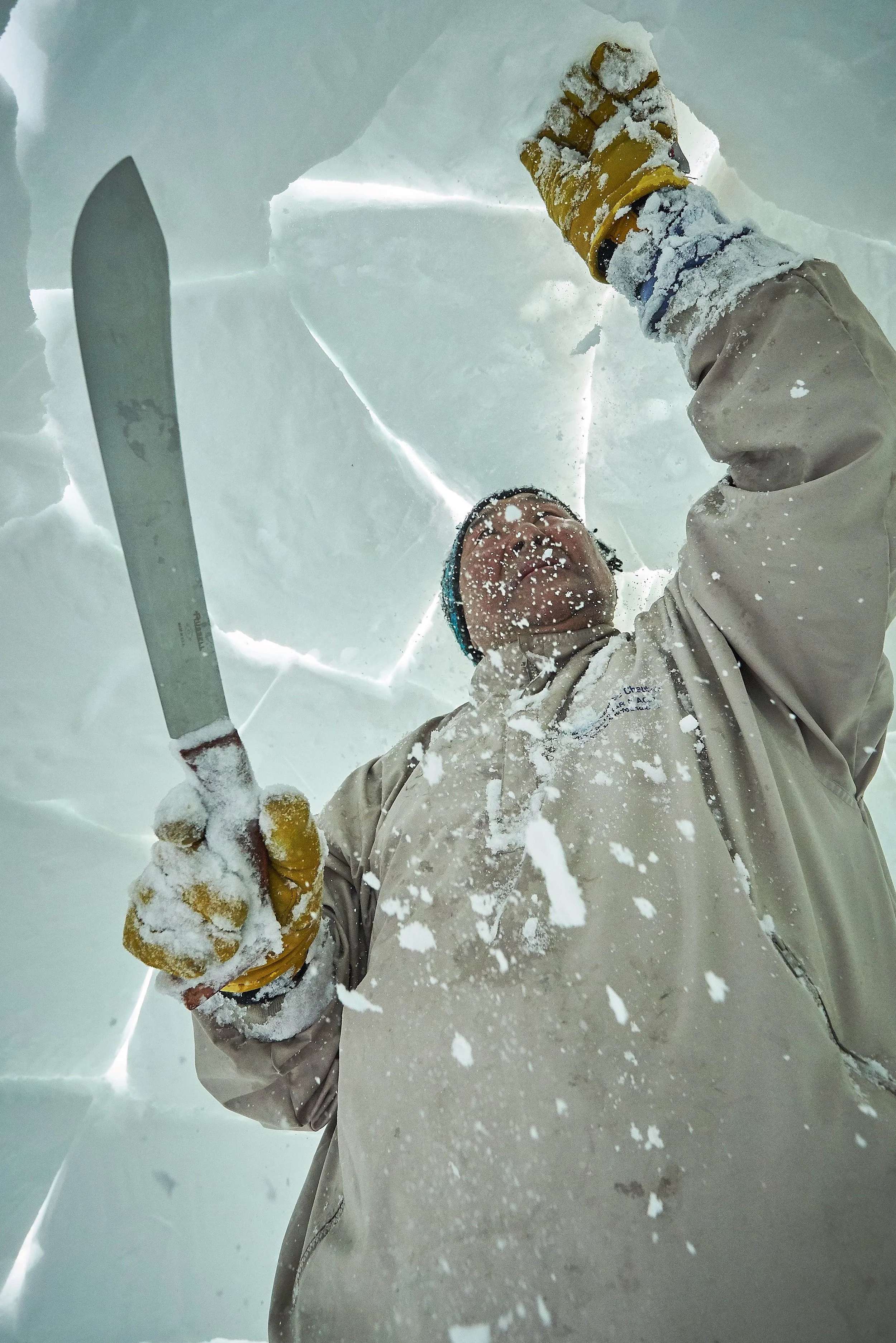 Person inside a large ice cave, wearing beige winter gear and yellow gloves, holding a knife, and looking upward, surrounded by icy walls.