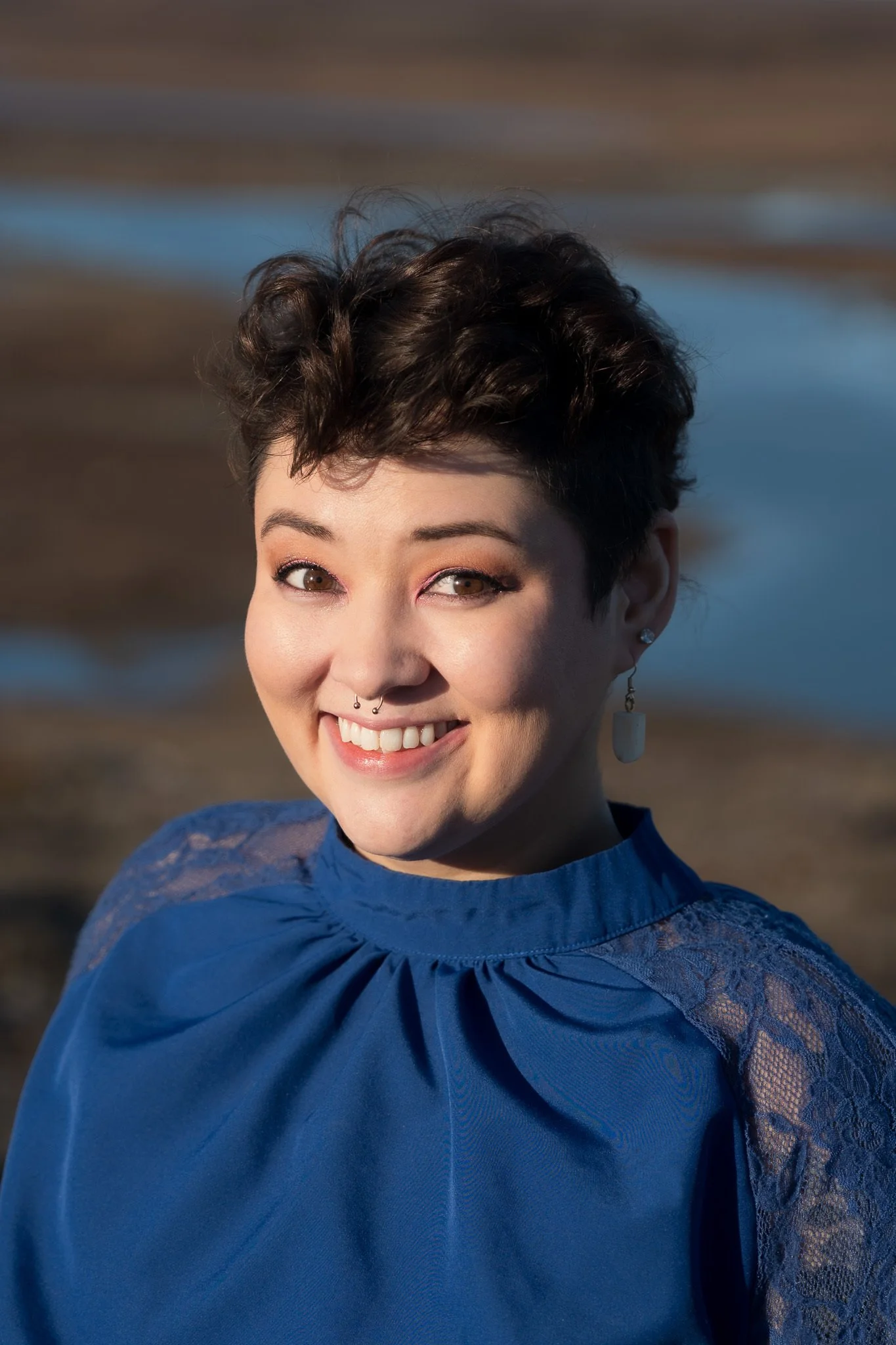 A woman with short dark hair smiling outdoors with a blurred background of water and land.