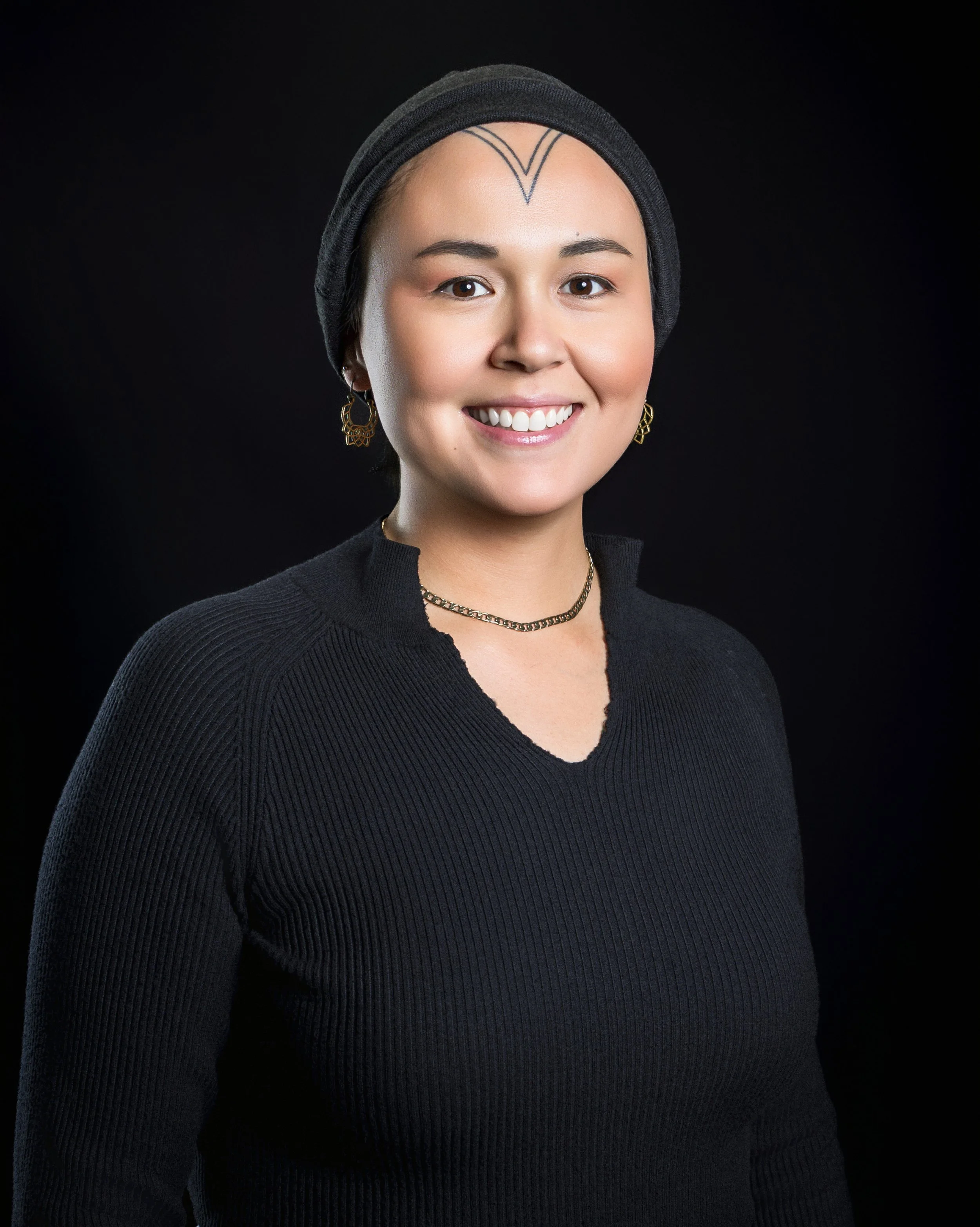 Portrait of a young woman smiling, wearing a black headscarf, black sweater, jewelry, and earrings against a black background.
