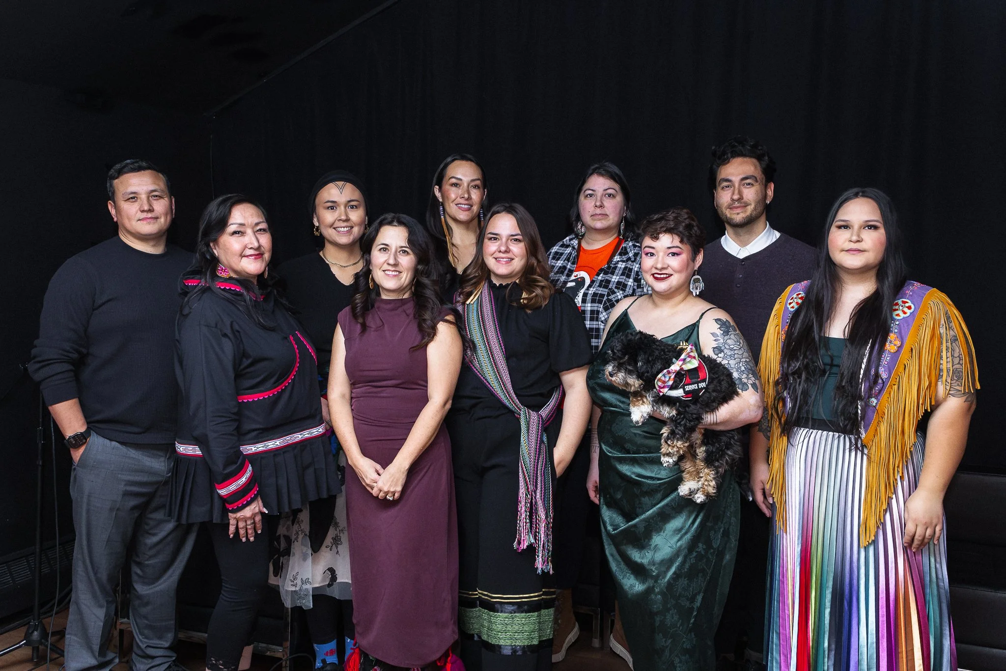 Group of people standing together, some in traditional clothing, with a woman holding a small dog. The background is black.