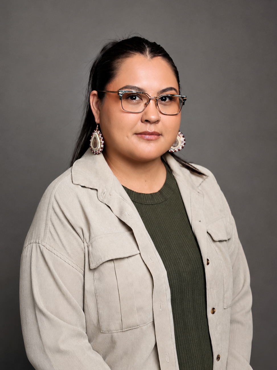 A woman with dark hair, glasses, and earrings, wearing a beige jacket over a dark green shirt, posed against a gray background.