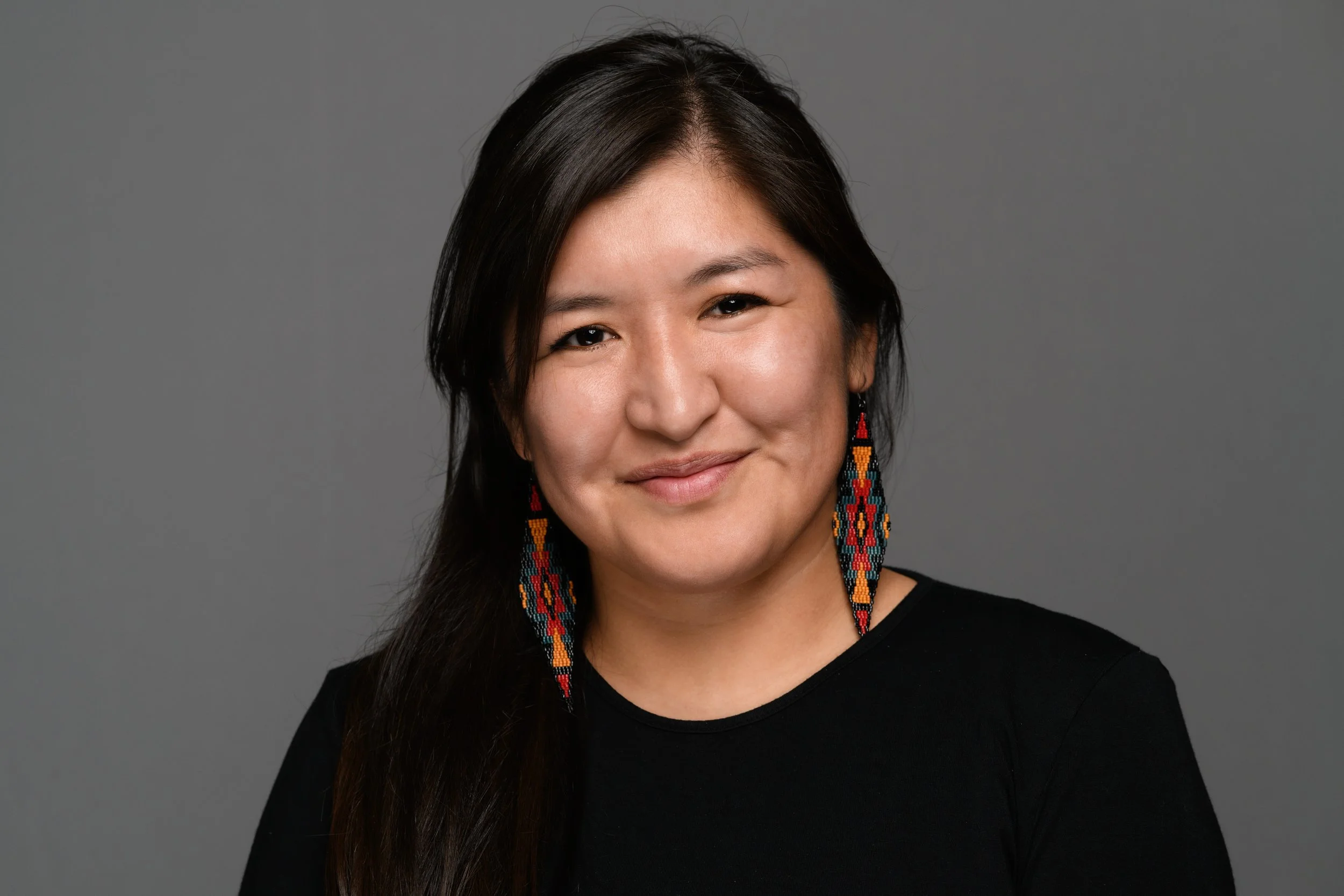 Portrait of a woman with long dark hair, earrings, and a black top, smiling against a gray background.