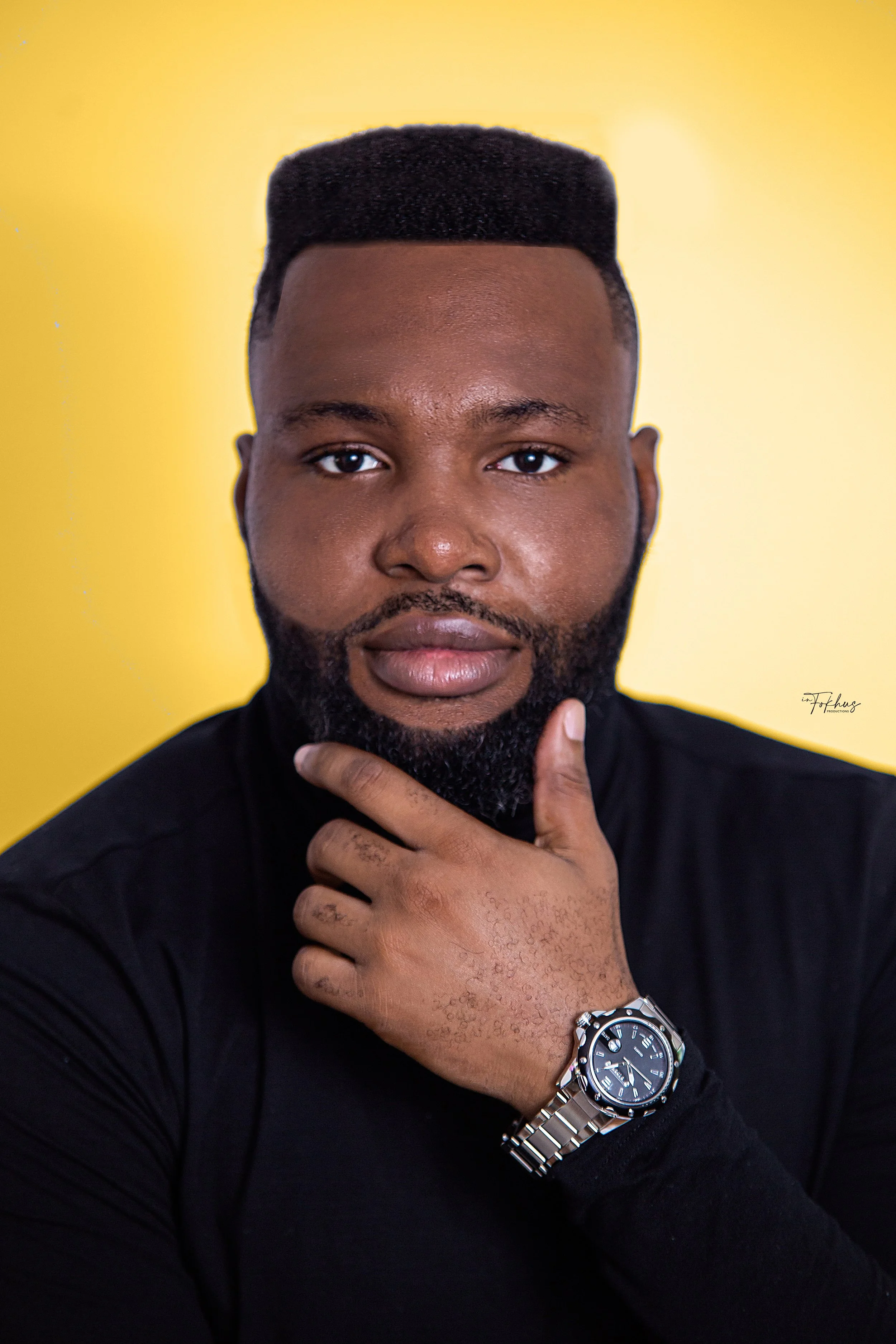 Portrait of a man with a short hairstyle and beard, wearing a black shirt, and a silver watch on his left wrist, against a yellow background.