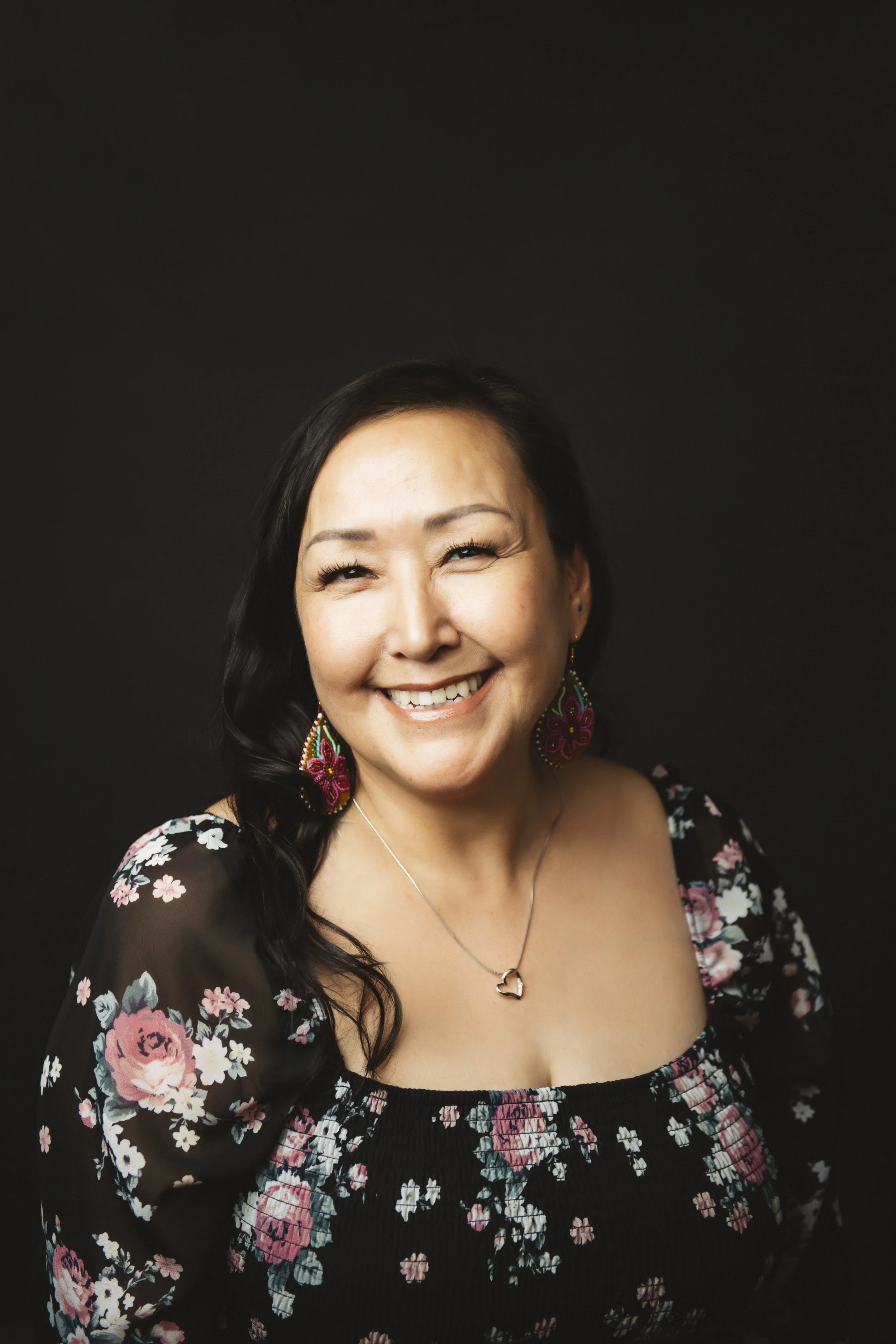 A smiling woman with dark hair, wearing floral earrings, a floral top, and a heart-shaped necklace, against a black background.