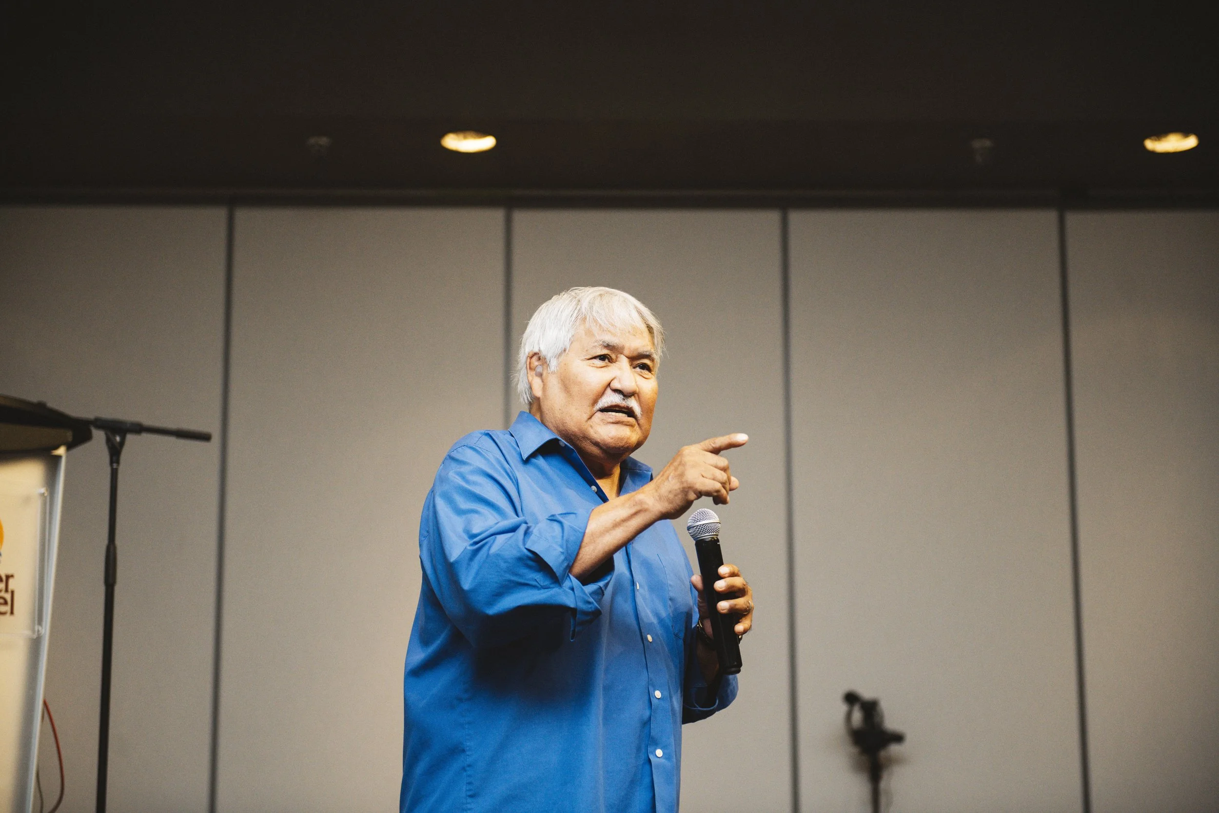 An elderly man with gray hair wearing a blue shirt, speaking into a microphone and gesturing with his right hand in a conference room.