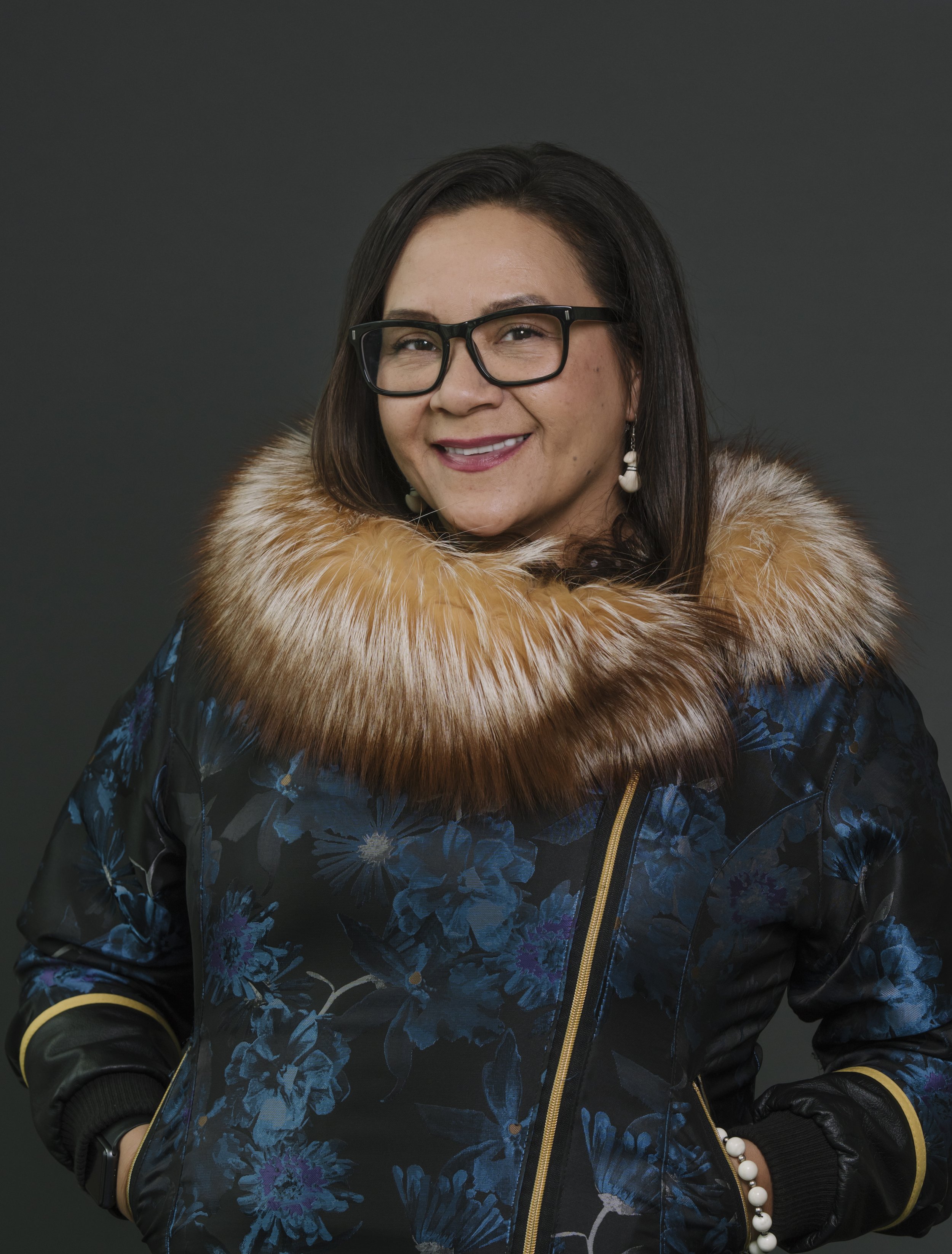 A woman with dark hair, wearing glasses, a floral patterned jacket with a fur collar, and earrings, smiling against a dark background.