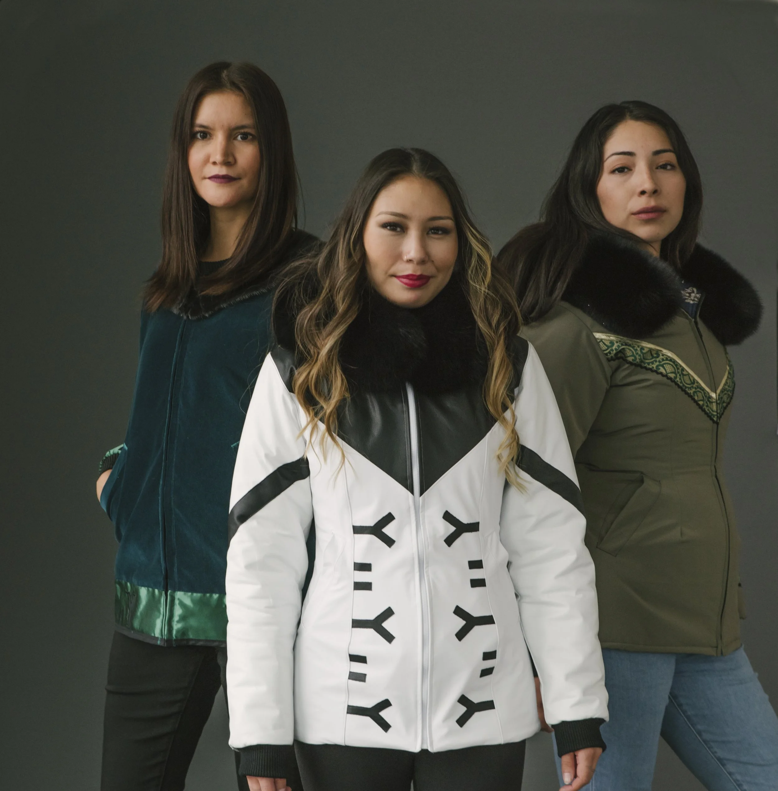 Three women standing together against a dark background, wearing stylish winter jackets with fur-lined hoods.