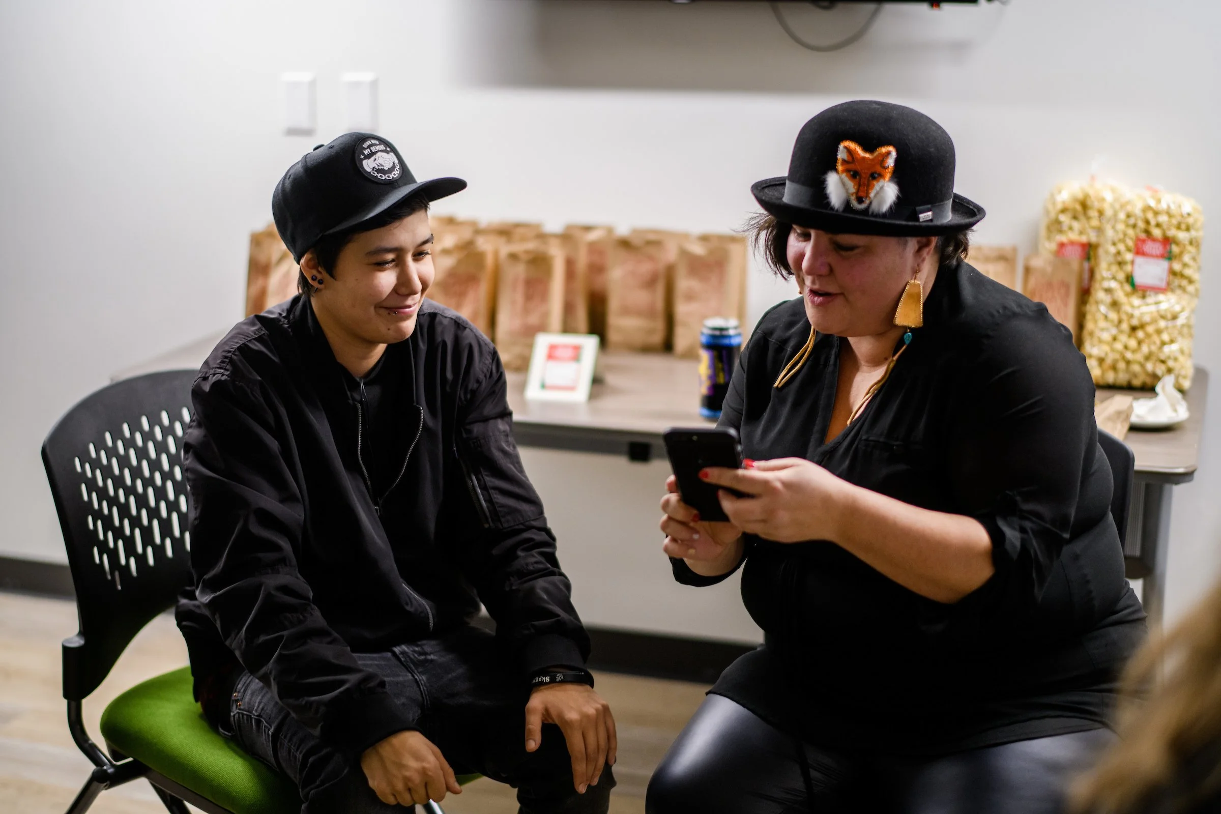 Two women sitting in a room, one wearing a black hat with a fox patch, checking her phone, the other wearing a black cap with a patch, smiling while sitting on a green chair.
