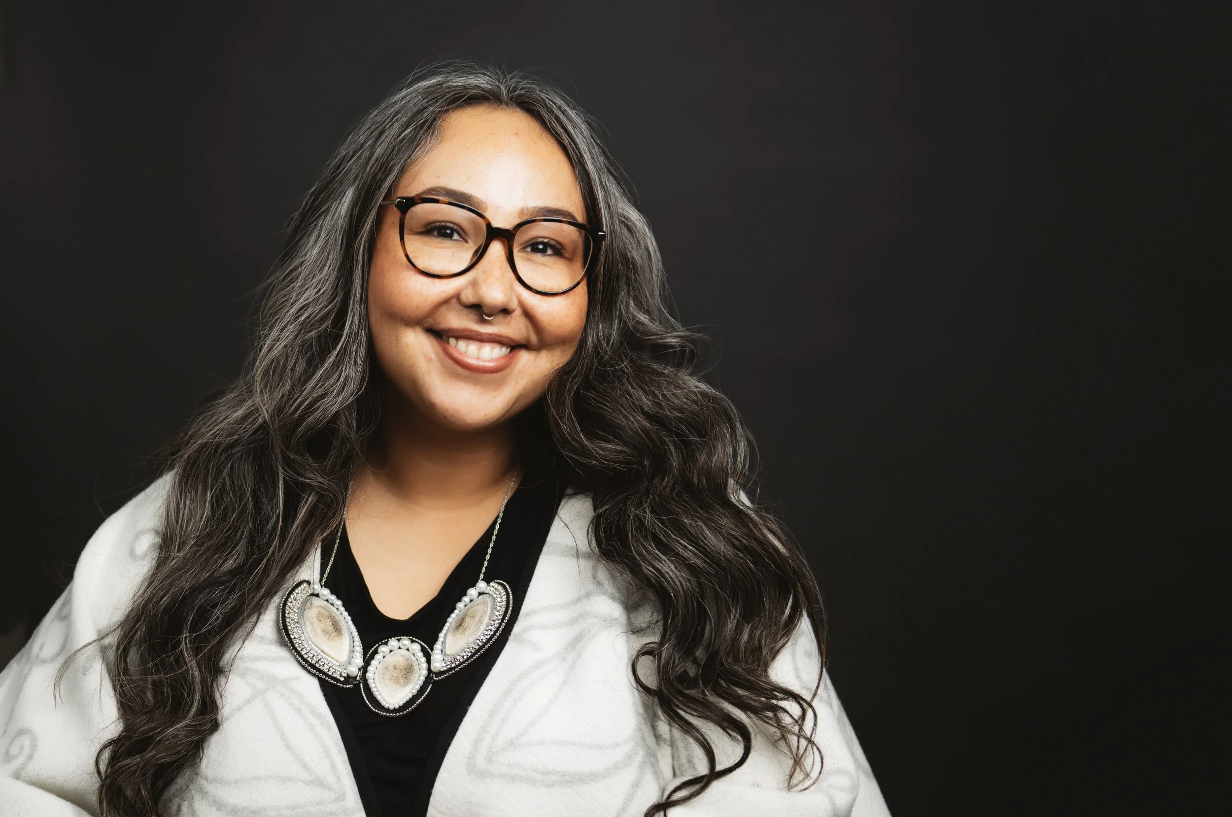 Smiling woman with long wavy hair, glasses, and a large necklace, wearing a white jacket, against a dark background.
