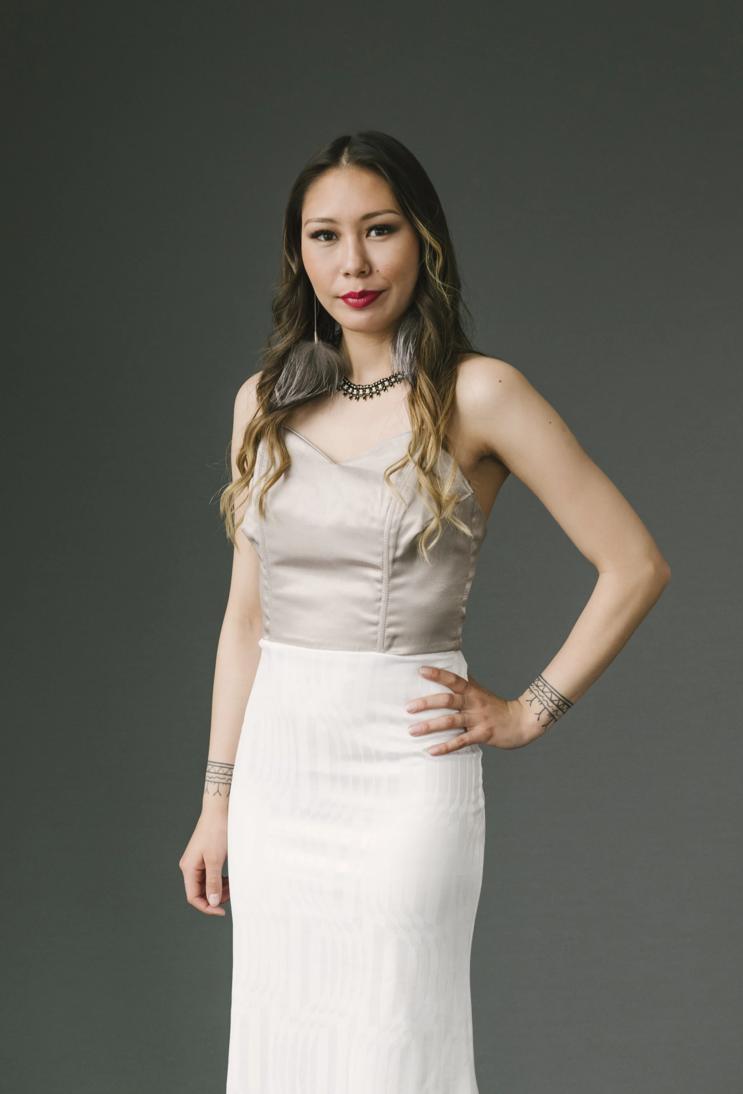 A woman with long wavy hair wearing a silver satin top, white skirt, and accessorized with a choker necklace and tattoos, standing against a plain gray background.