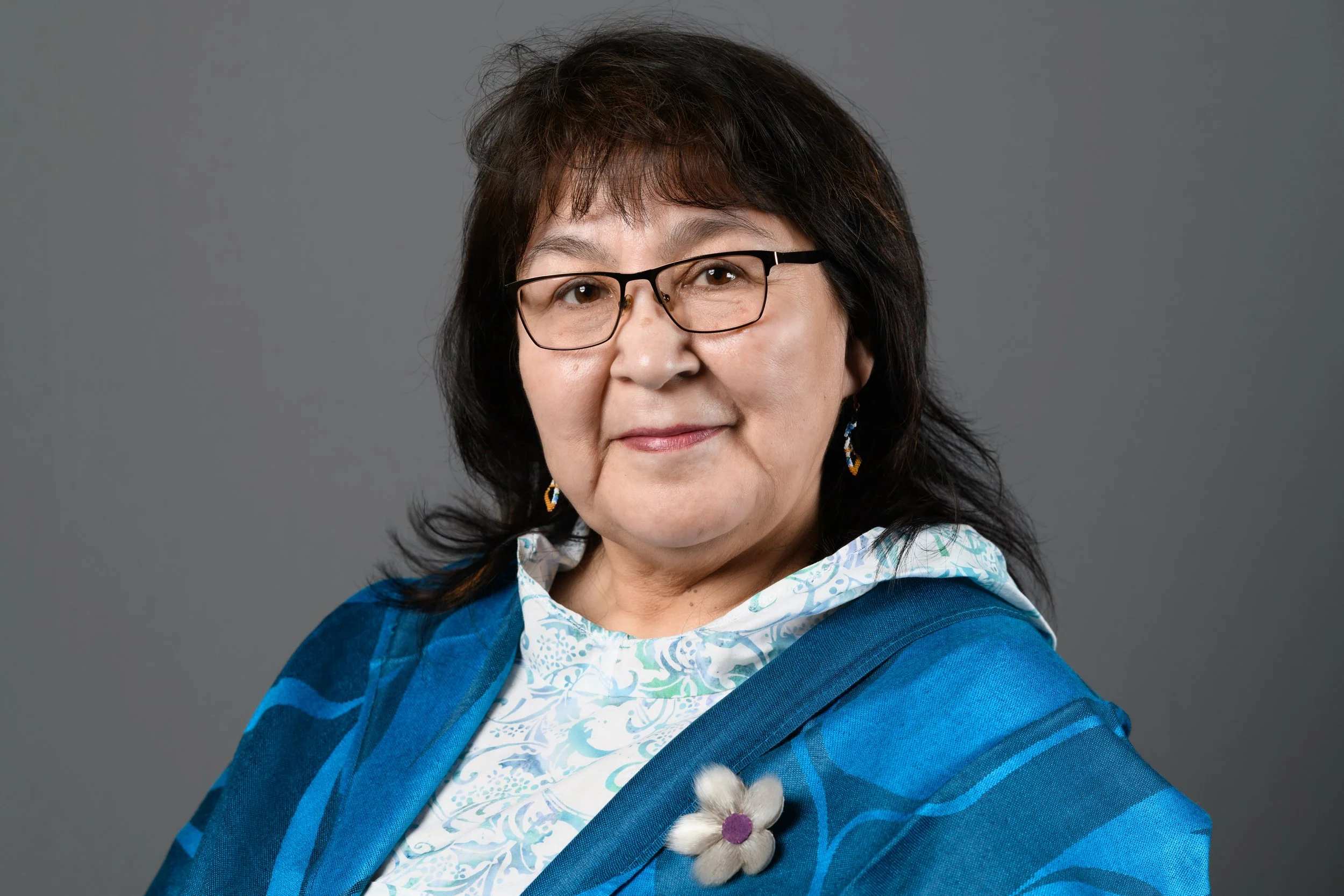 Portrait of an older woman with glasses, black hair, wearing a blue patterned jacket and white blouse with a flower pin, against a gray background.