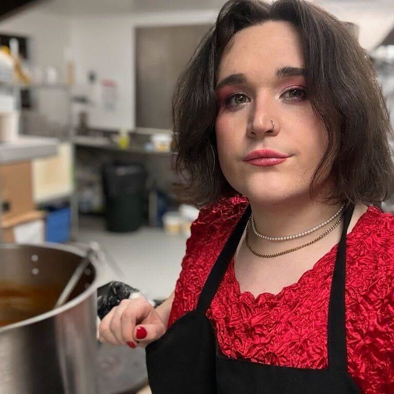 A woman with shoulder-length dark hair and light skin wearing a red top, black apron, and jewelry, standing in a kitchen with various utensils and containers in the background.