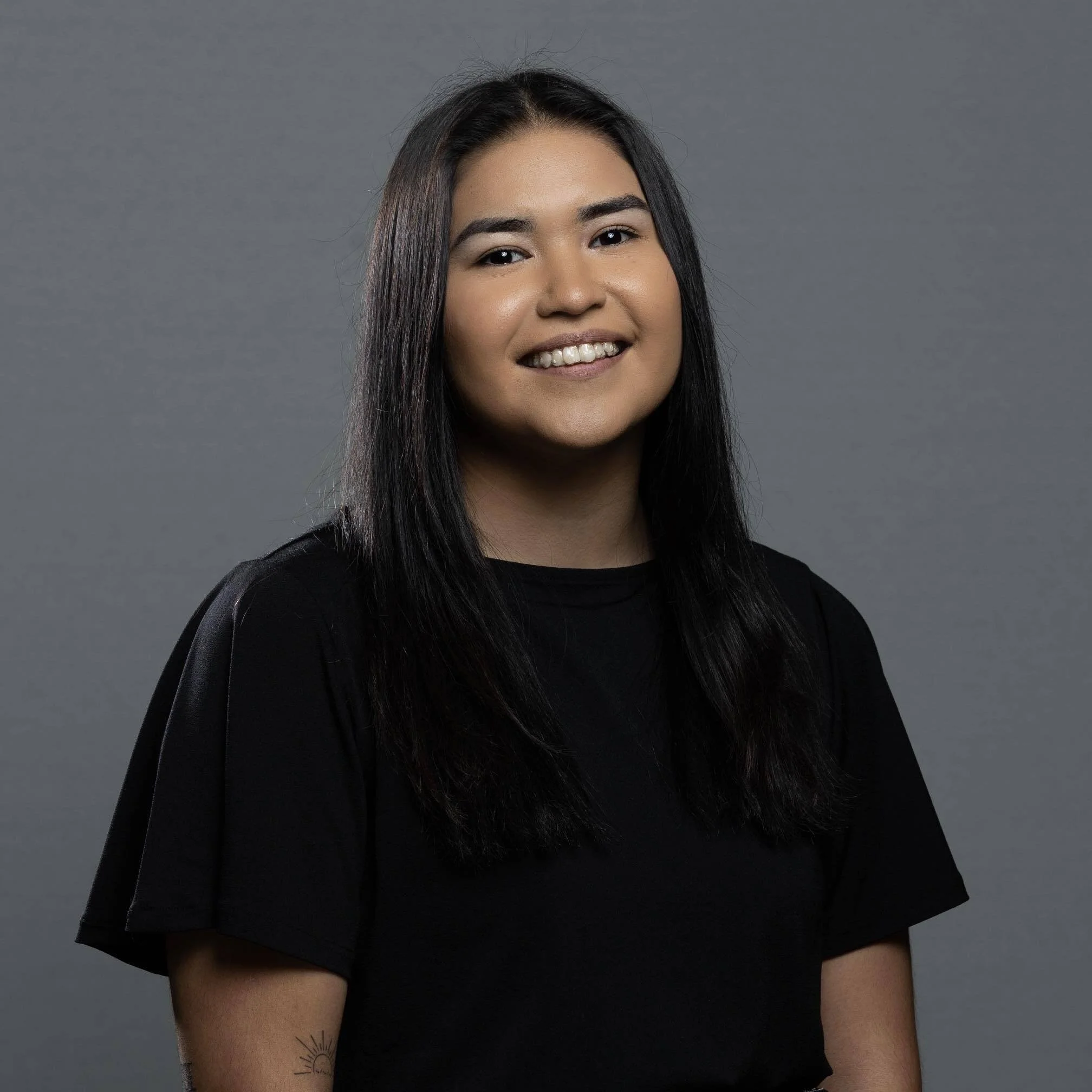 Portrait of a young woman with long black hair, smiling, against a gray background.