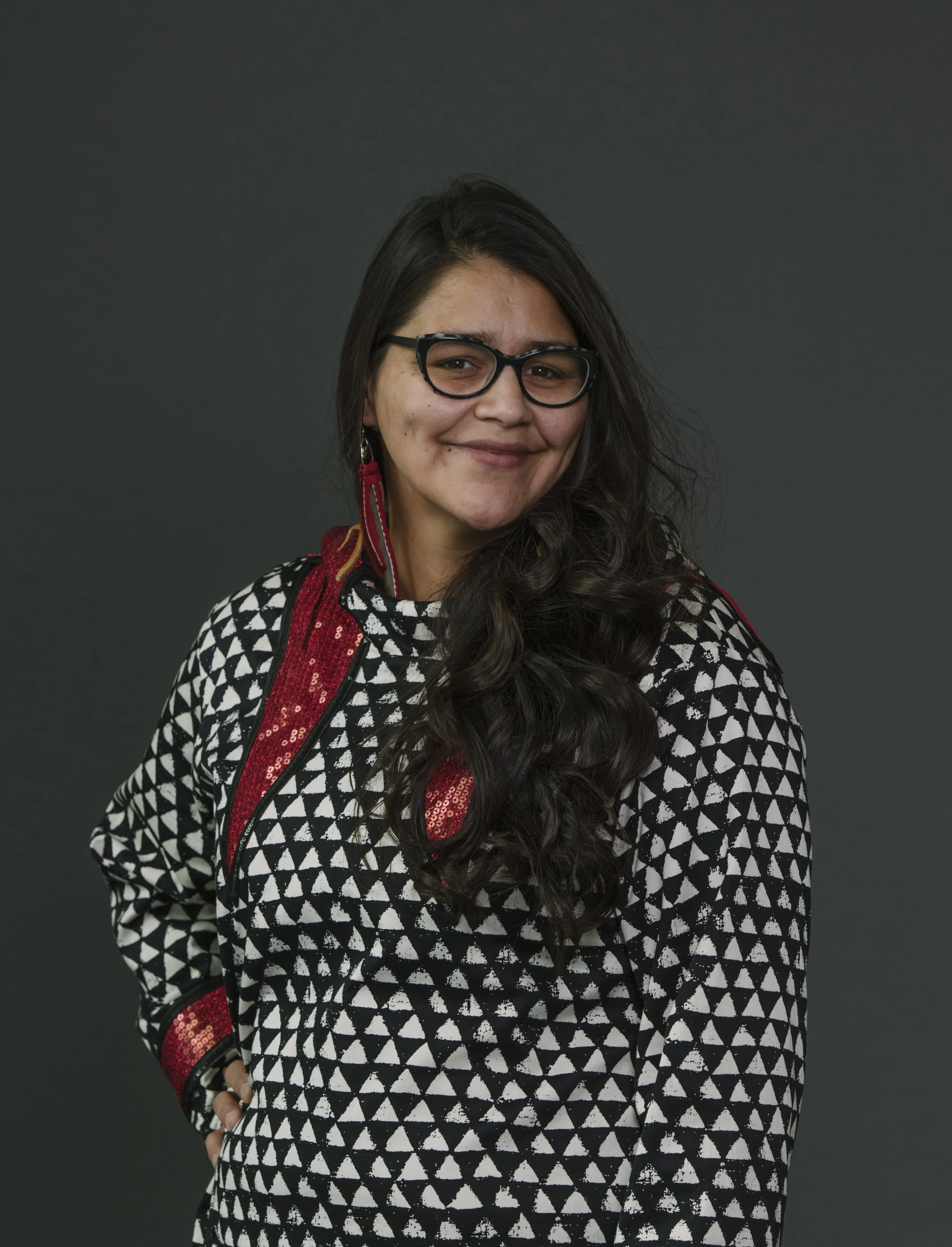 A woman with long dark hair, glasses, and earrings, smiling, wearing a black and white patterned top with red accents, standing against a dark background.