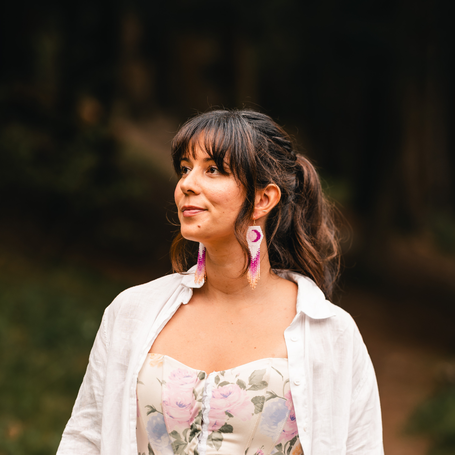 A woman with dark brown, wavy hair tied in a ponytail, wearing long pink and purple beaded earrings, a white open-button shirt, and a floral dress, standing outdoors with a blurred nature background.
