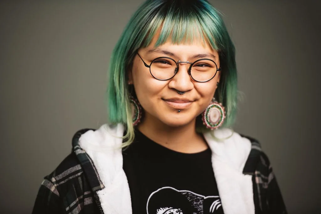 Portrait of a woman with green hair, glasses, large earrings, and facial piercings, smiling at the camera.