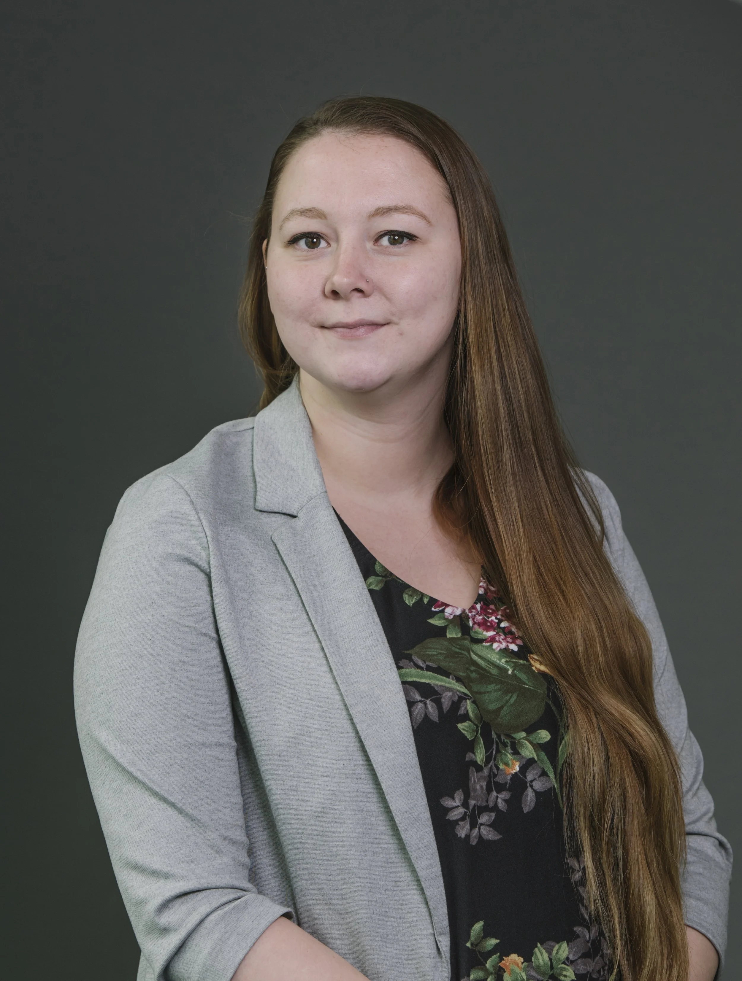 Portrait of a young woman with long brown hair, wearing a gray blazer over a black floral top, standing against a dark gray background.