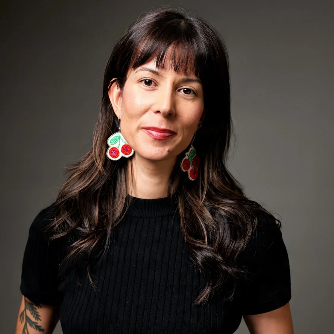 A woman with dark brown, wavy hair tied in a ponytail, wearing long pink and purple beaded earrings, a white open-button shirt, and a floral dress, standing outdoors with a blurred nature background.