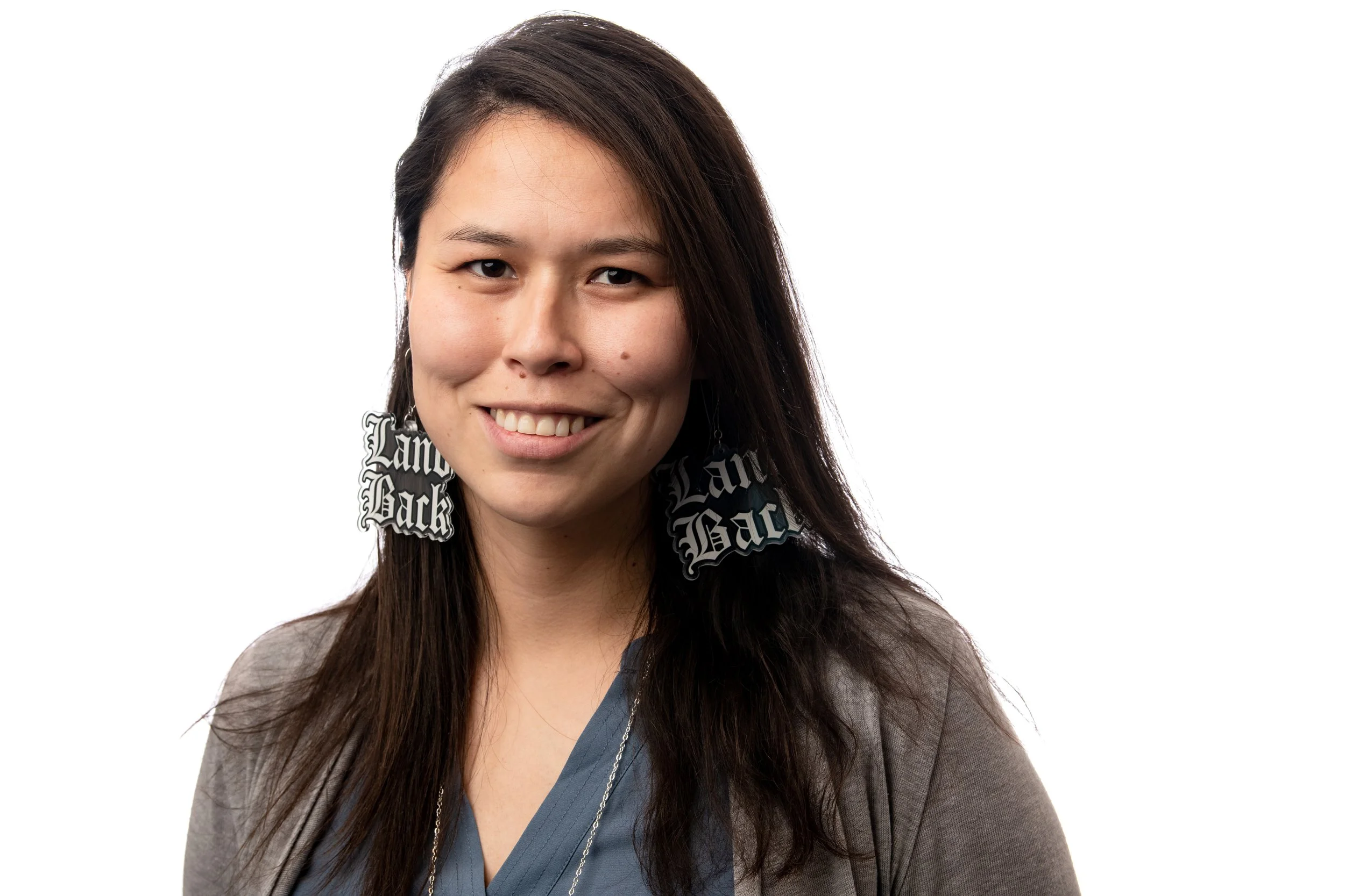 A young woman with long dark hair smiling, wearing large earrings that say "Land Back" and a gray cardigan over a blue top, against a plain white background.