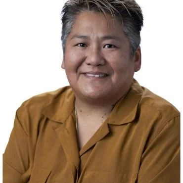 Portrait of a smiling person with short gray hair wearing a mustard yellow shirt.