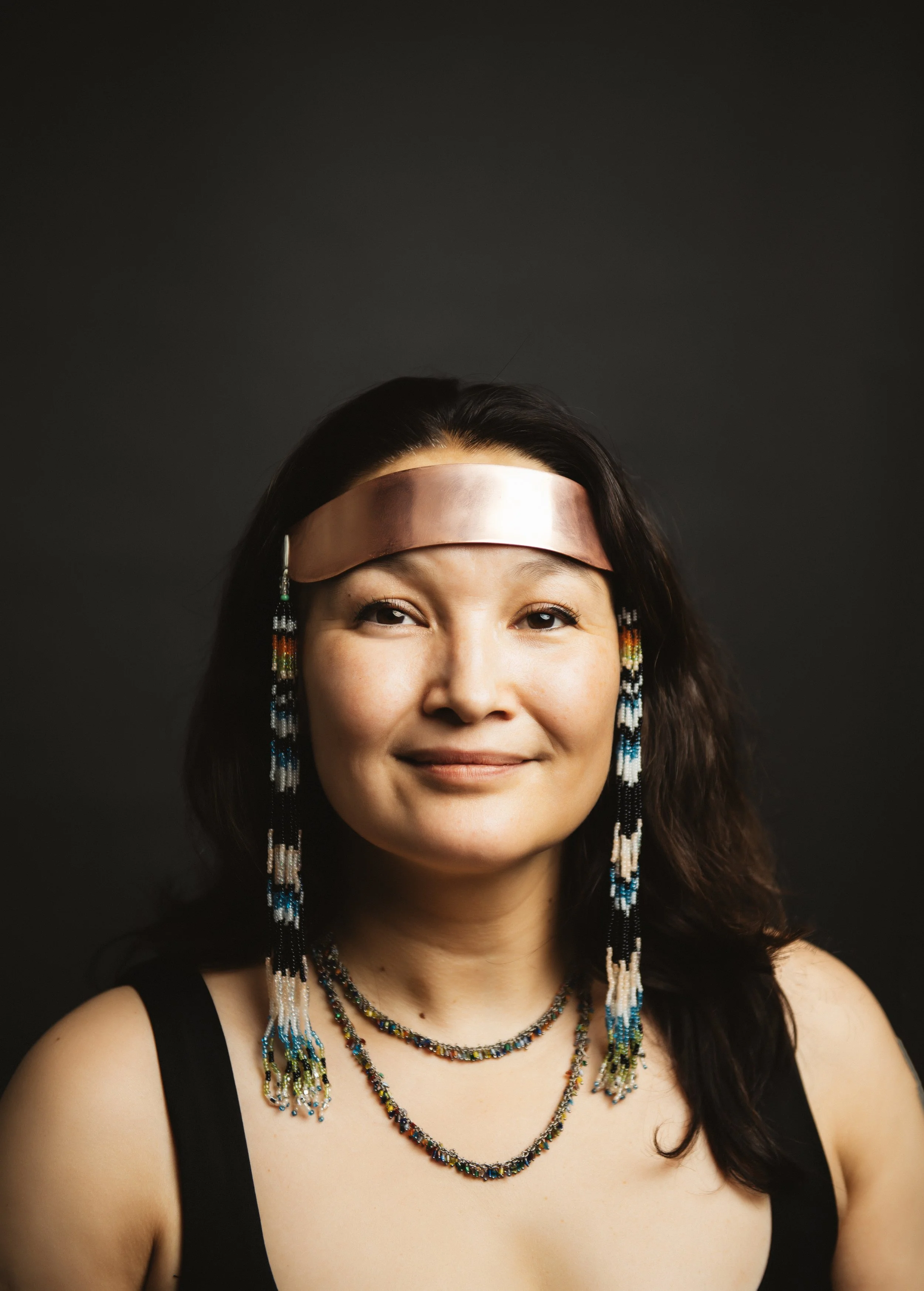 Portrait of a woman with long dark hair, wearing beaded jewelry and a metallic headband, against a dark background.
