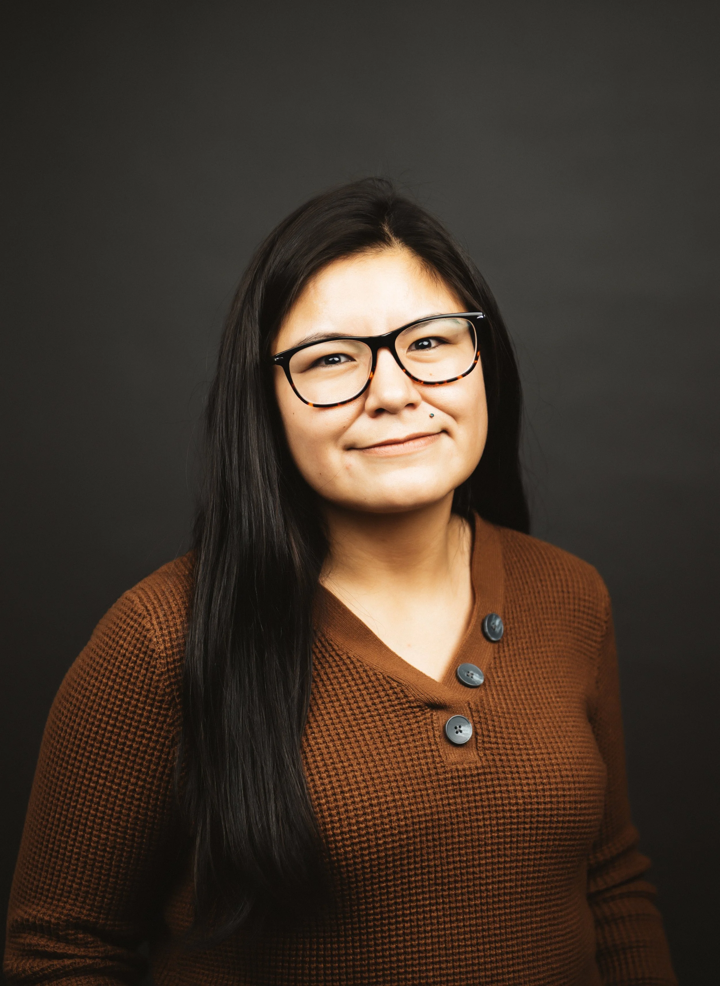 Woman with long black hair, glasses, and a brown textured top standing against a dark background.