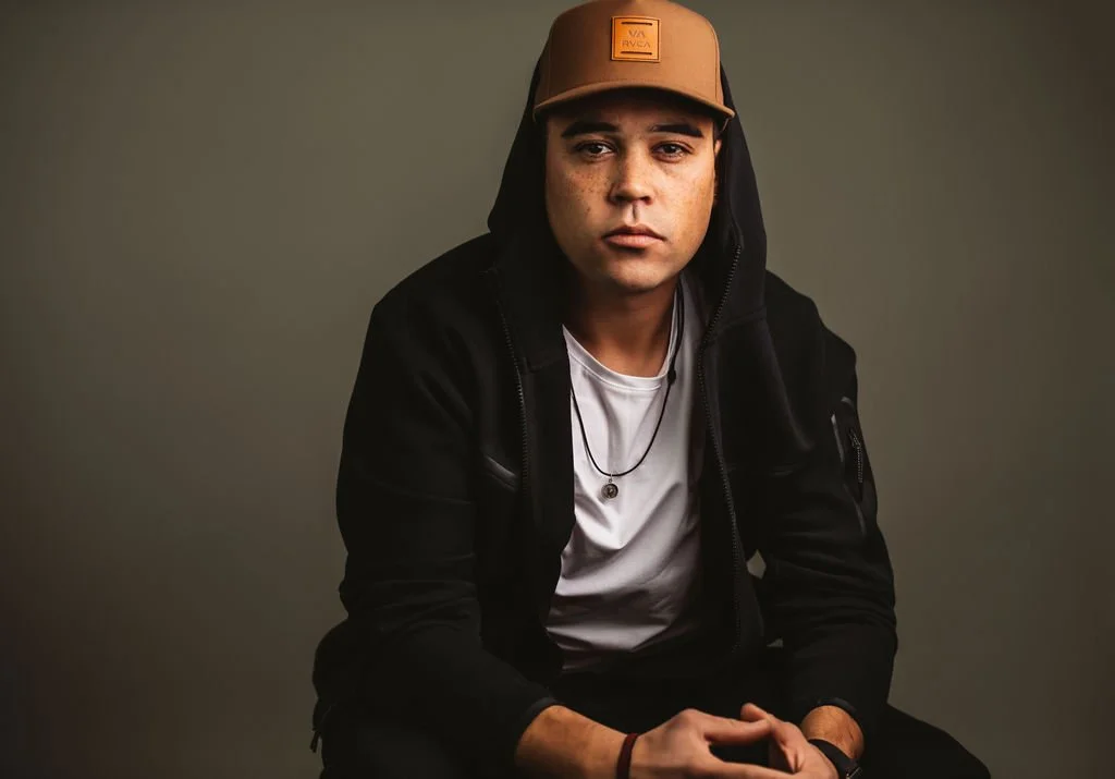 A young man wearing a beige cap, black hoodie, and white t-shirt sitting against a plain background.
