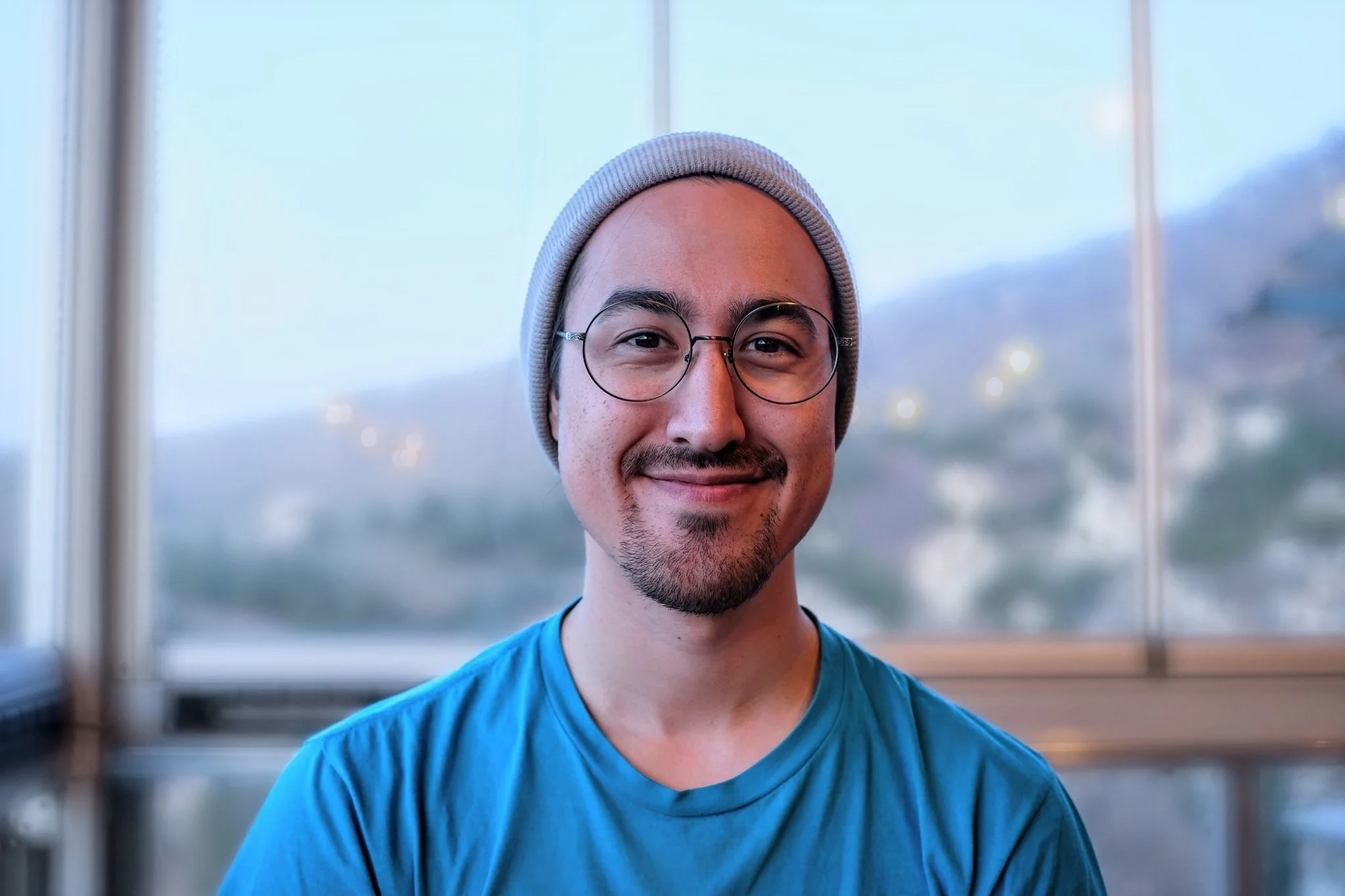 A young man smiling, wearing glasses, a gray beanie, and a blue t-shirt, sitting in front of a large window with a scenic mountain backdrop.