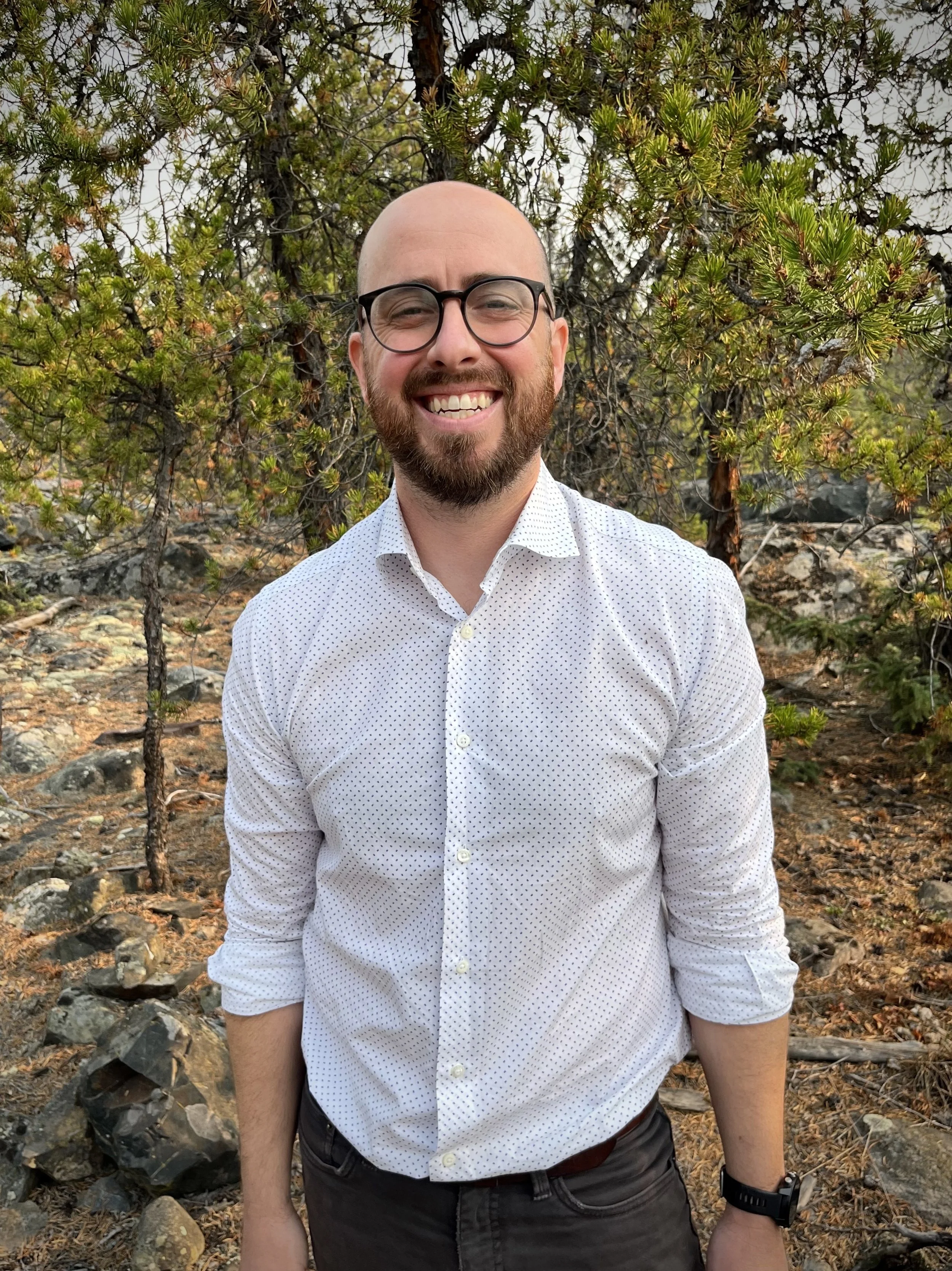 A smiling man with a beard and glasses, wearing a white dotted shirt, standing outdoors among trees and rocky terrain.