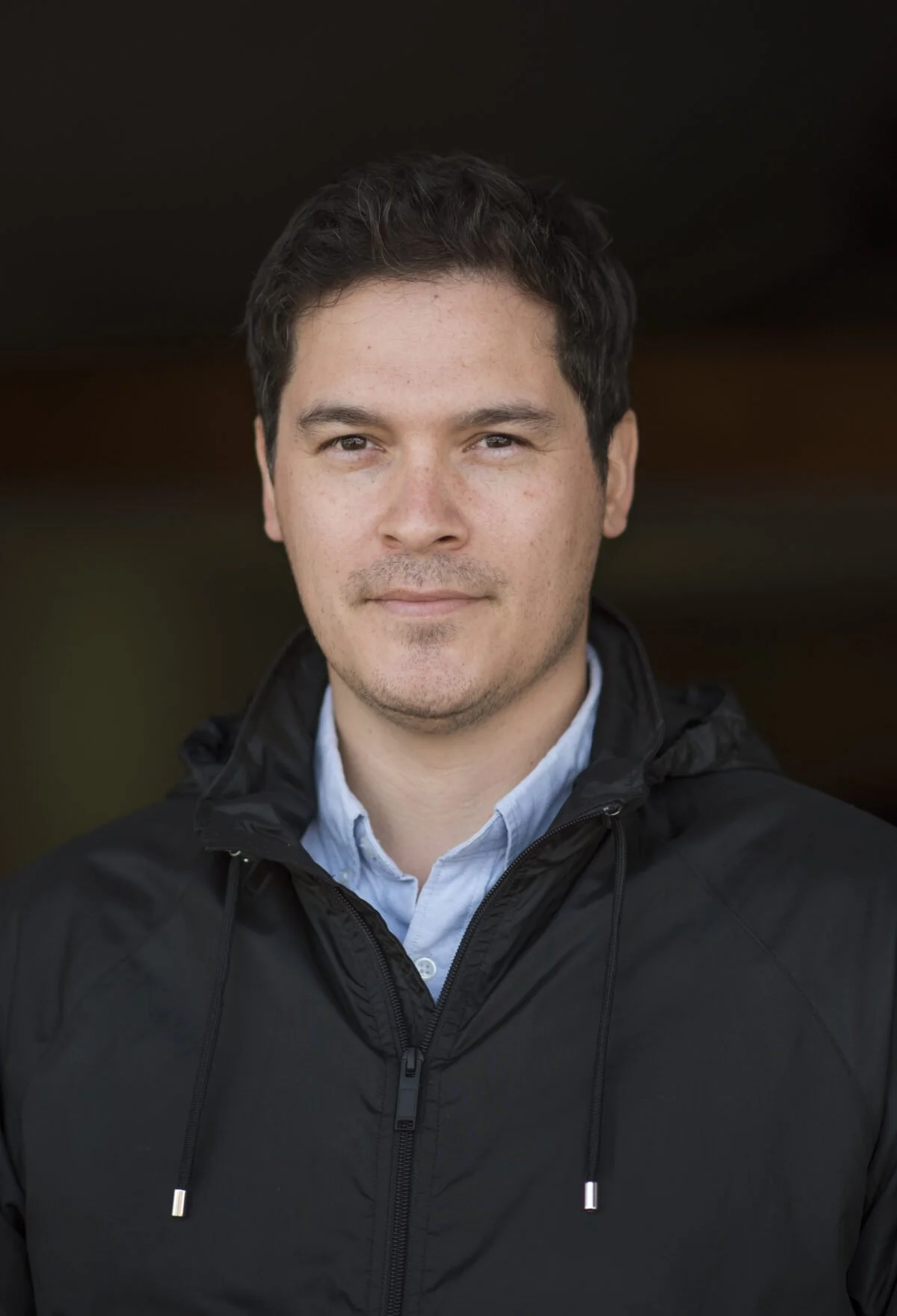 A portrait of a young man with dark hair, light skin, wearing a black jacket and a blue collared shirt, standing outdoors with a dark background.