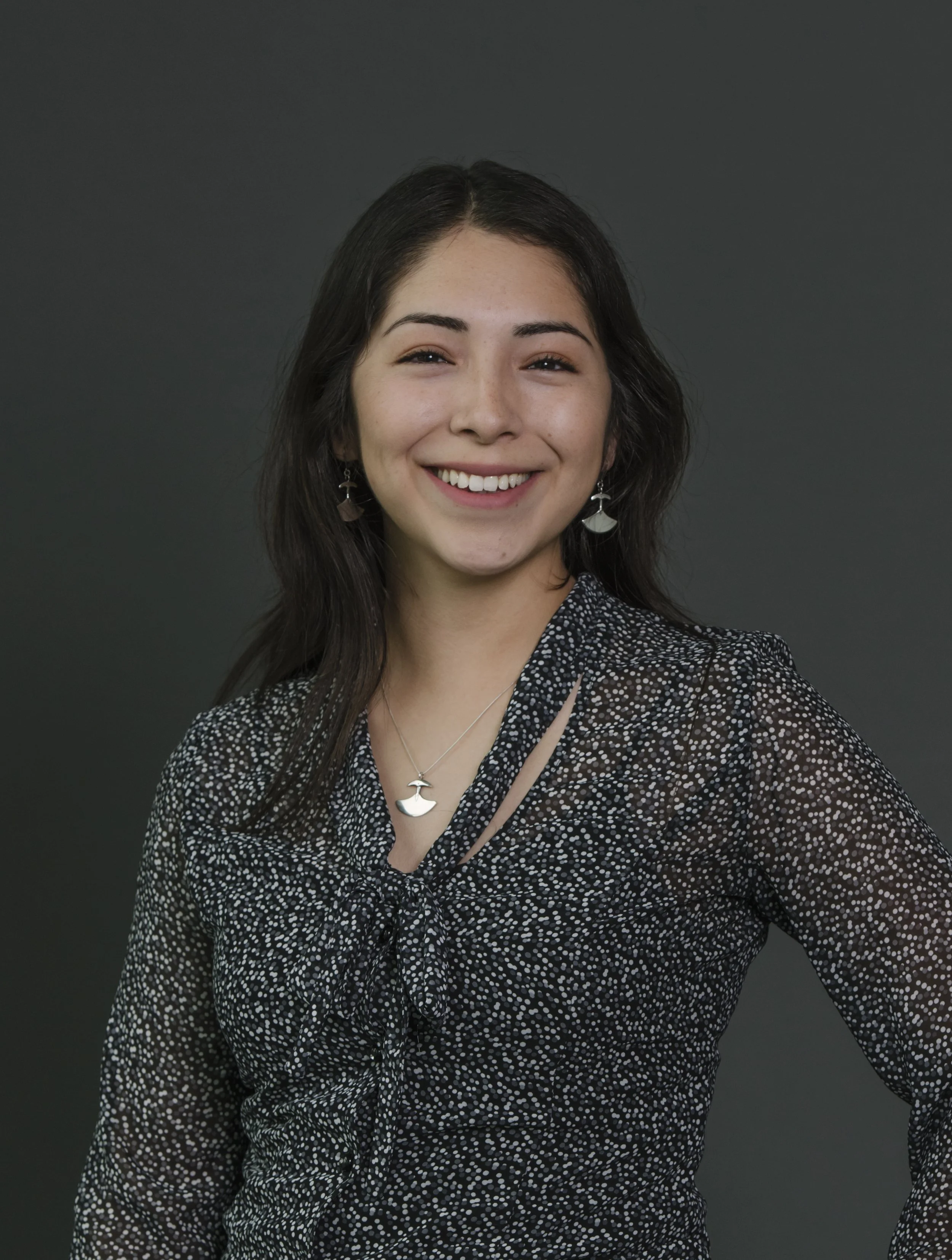A young woman with long dark hair, smiling, wearing jewelry including earrings and a necklace with a moon-shaped pendant, and a black and white polka-dot dress with a bow tie detail.