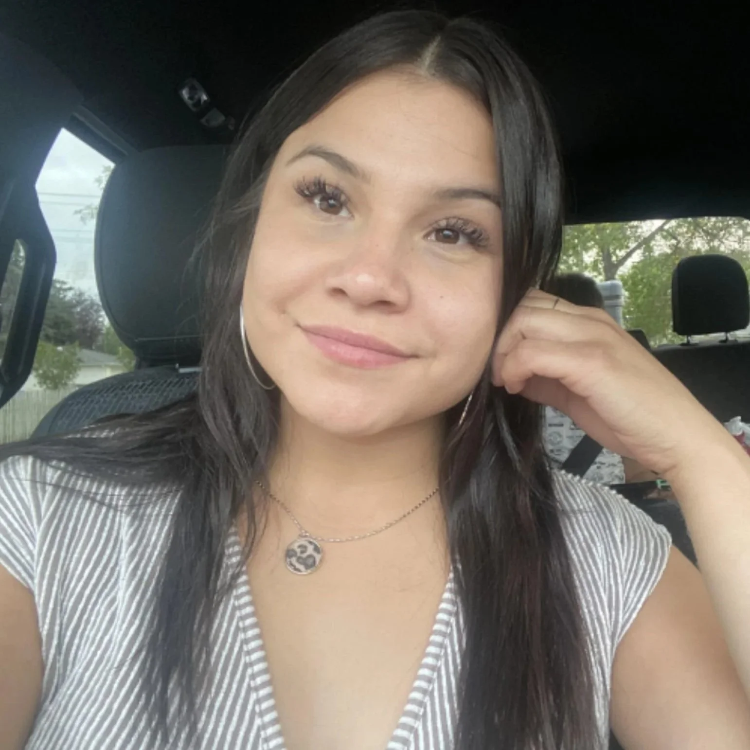 A young woman with long dark hair, wearing hoop earrings, a necklace with a yin-yang pendant, and a striped shirt, smiling while sitting in a vehicle.
