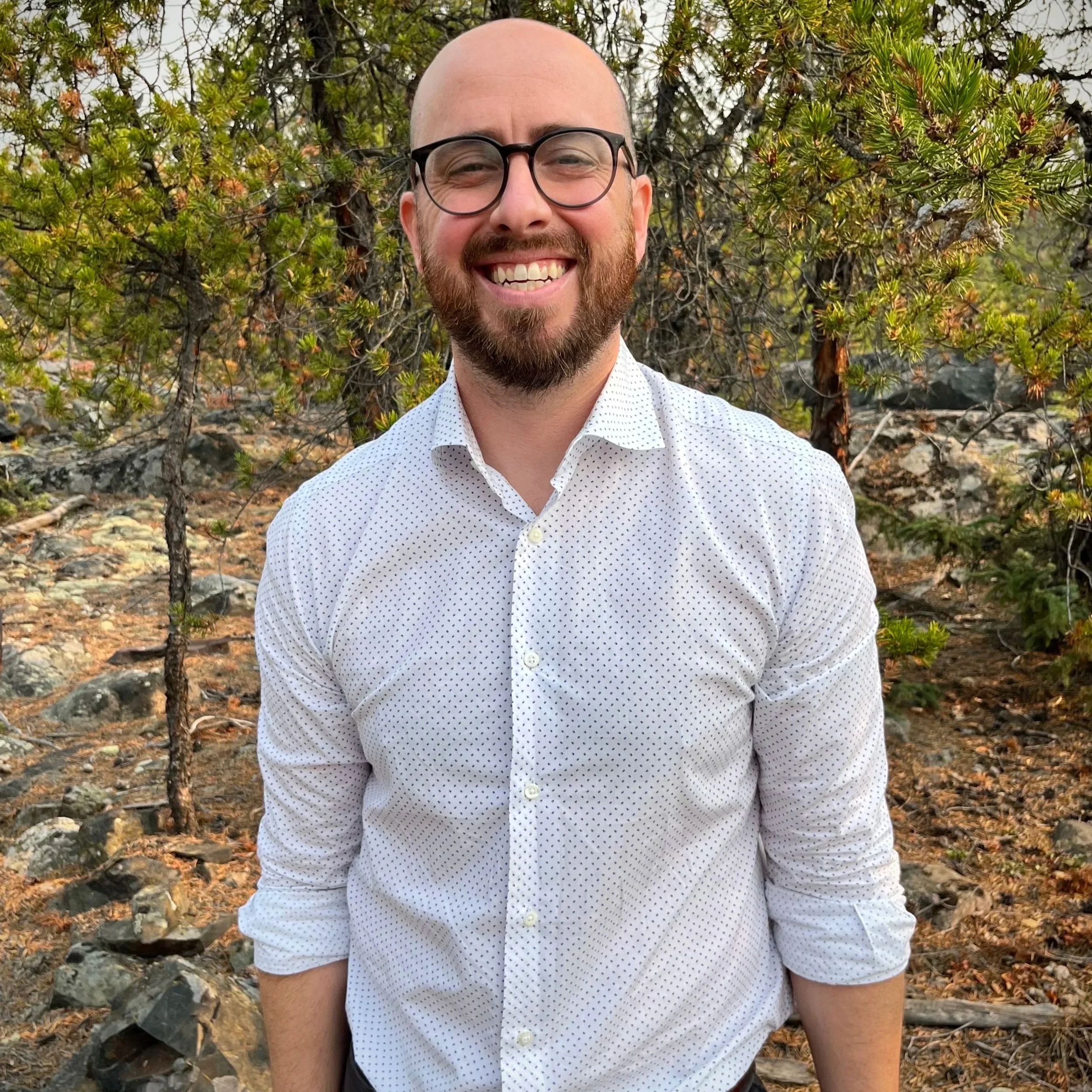 Smiling man with glasses and a beard, wearing a white shirt with small dark polka dots, standing outdoors among trees and rocks.