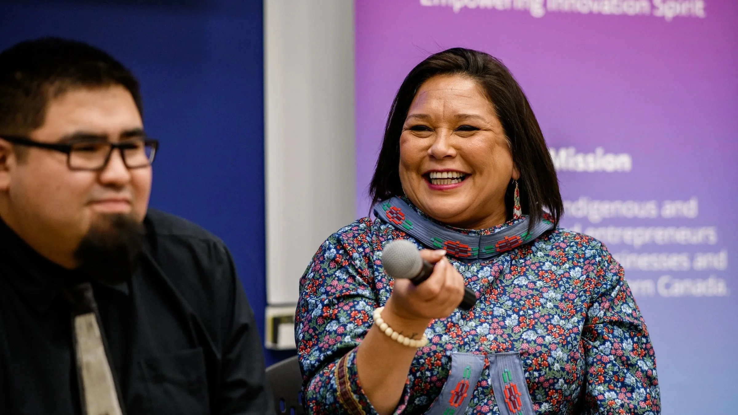 A woman in a colorful floral top holding a microphone and smiling at an indoor event, with a purple background and a man sitting next to her.