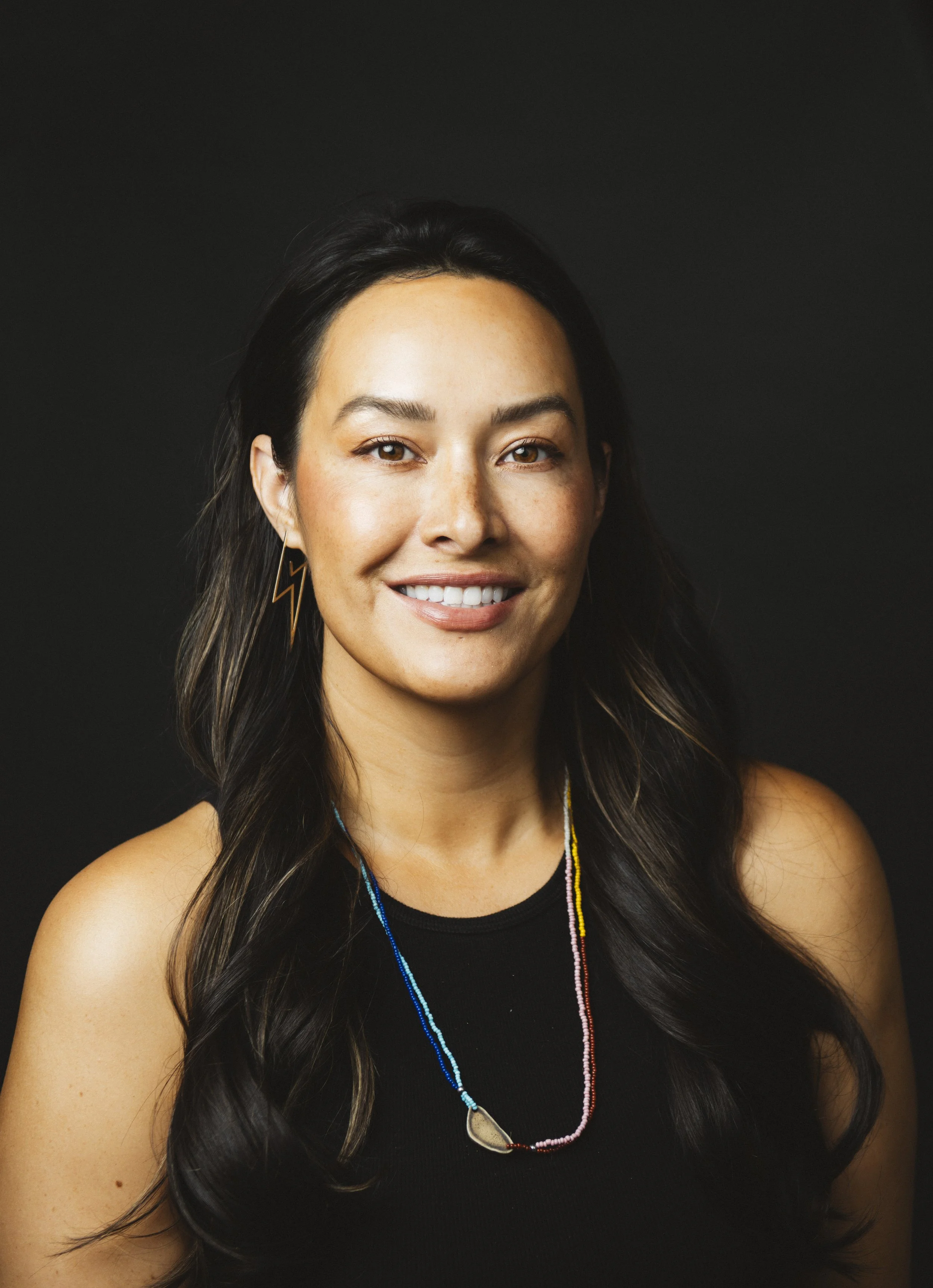 Portrait of a woman with long dark hair, wearing a black sleeveless top, colorful beaded necklace, and lightning bolt earrings, smiling against a dark background.