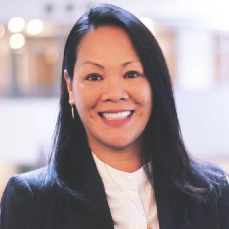 A woman with long black hair, wearing a dark blazer and white blouse, smiling in an indoor setting with blurred lights in the background.