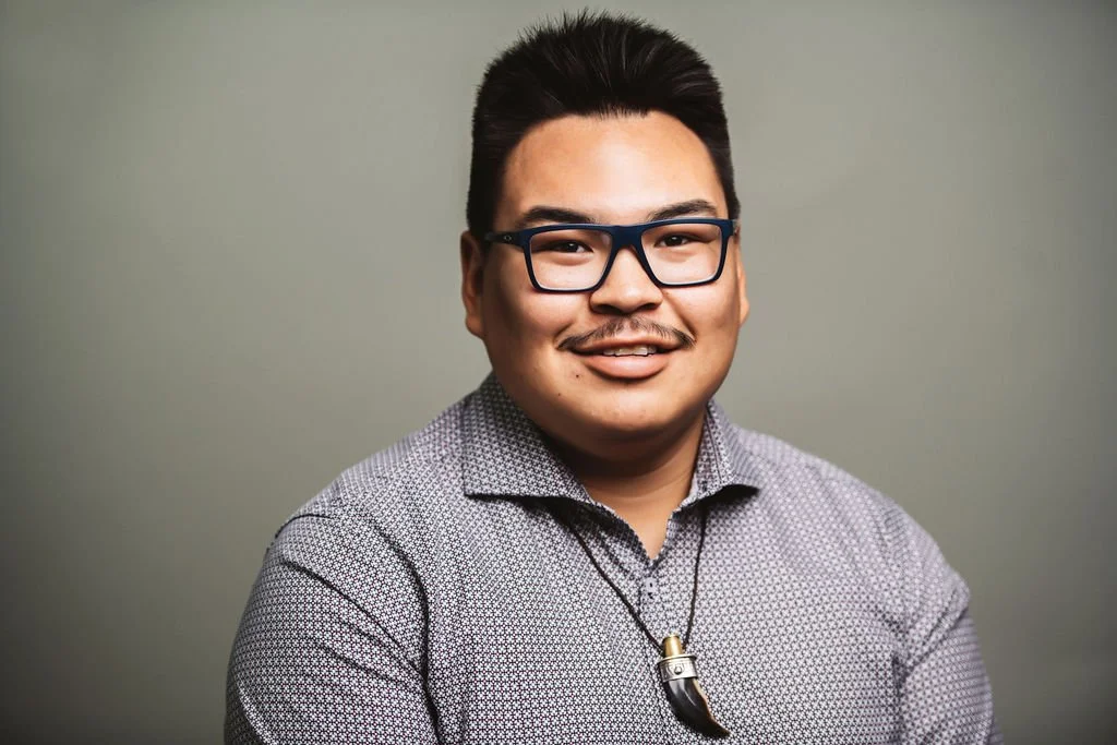 A smiling man with dark hair, wearing glasses, a patterned shirt, and a necklace with a pendant, standing against a plain background.
